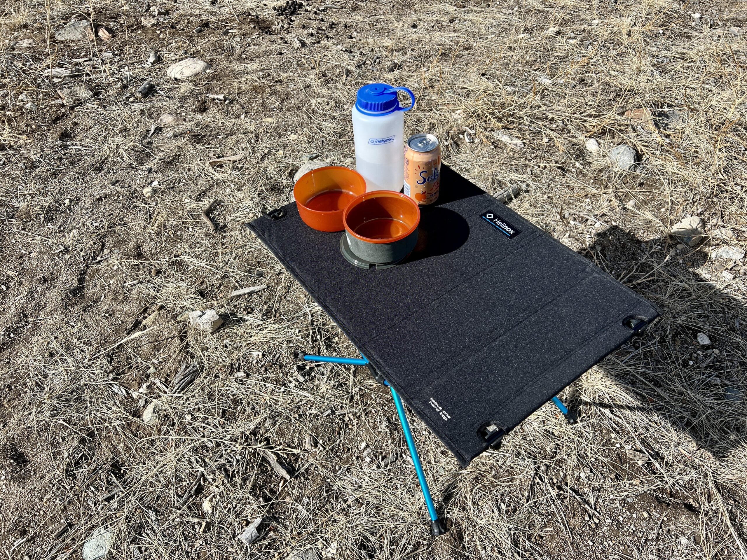 The Helinox One Hard Top Table stands on scrub and dirt. It has plasticware, a Nalgene, and a can of water on top.
