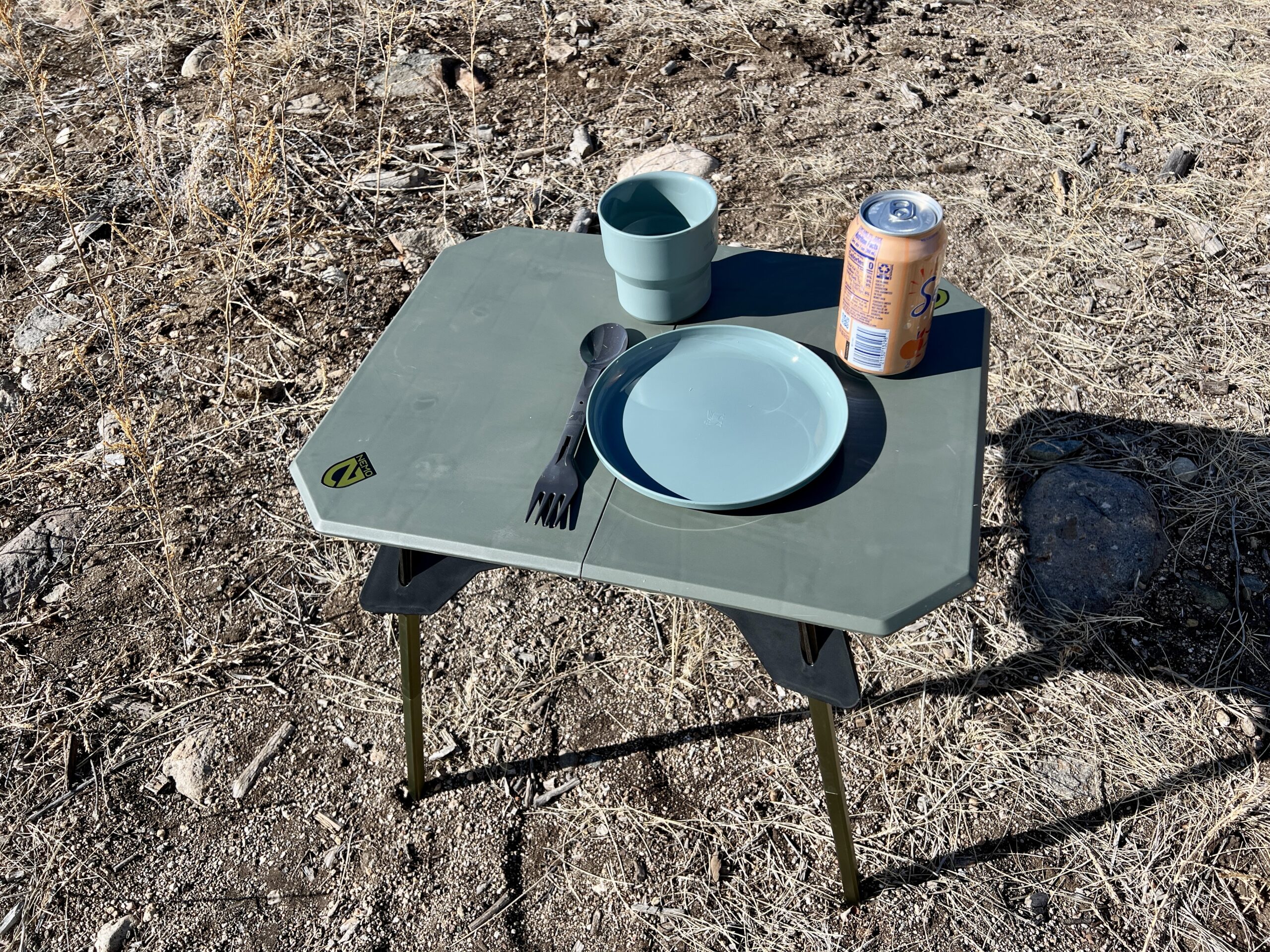 The NEMO Moonlander Dual-Height Table is standing on dirt with a can of water, a plate, a utensil, and a cup sitting on its top.