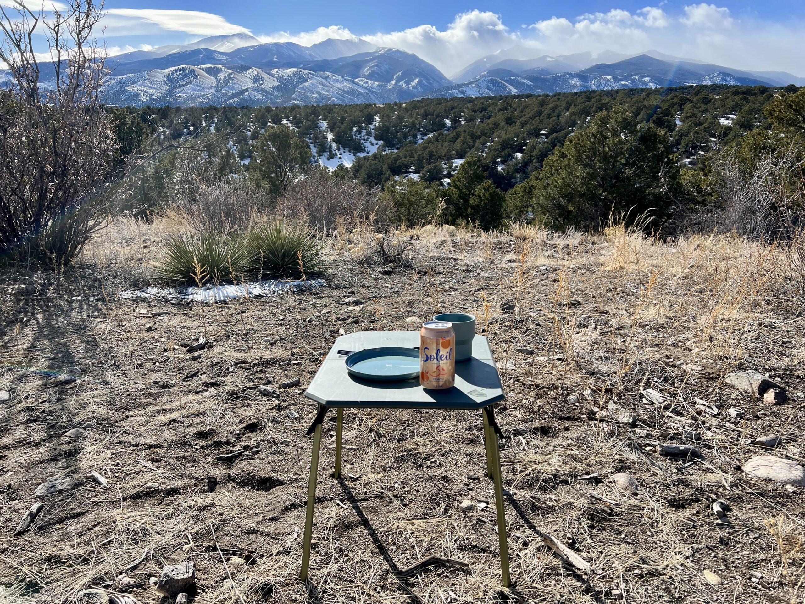 The NEMO Moonlander Dual-Height Camp Table stands outside with a mountain scene in the background. On its top, there is a can of water, a plate, and a cup.