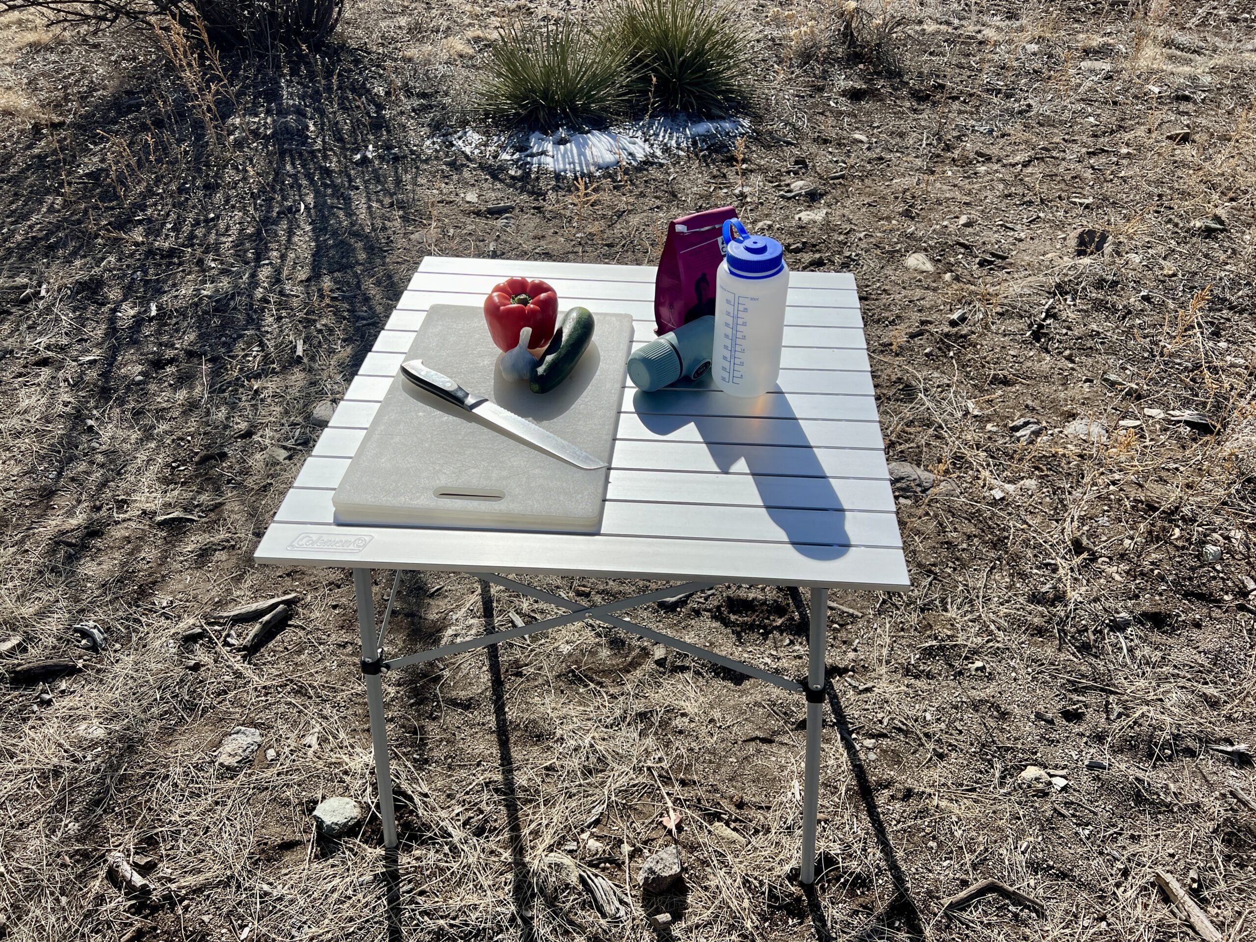 The Coleman Compact Roll-Top Table stands on dirt and has a cutting board, red pepper, zucchini, garlic, a Nalgene bottle, coffee, and Aeropress on its top.