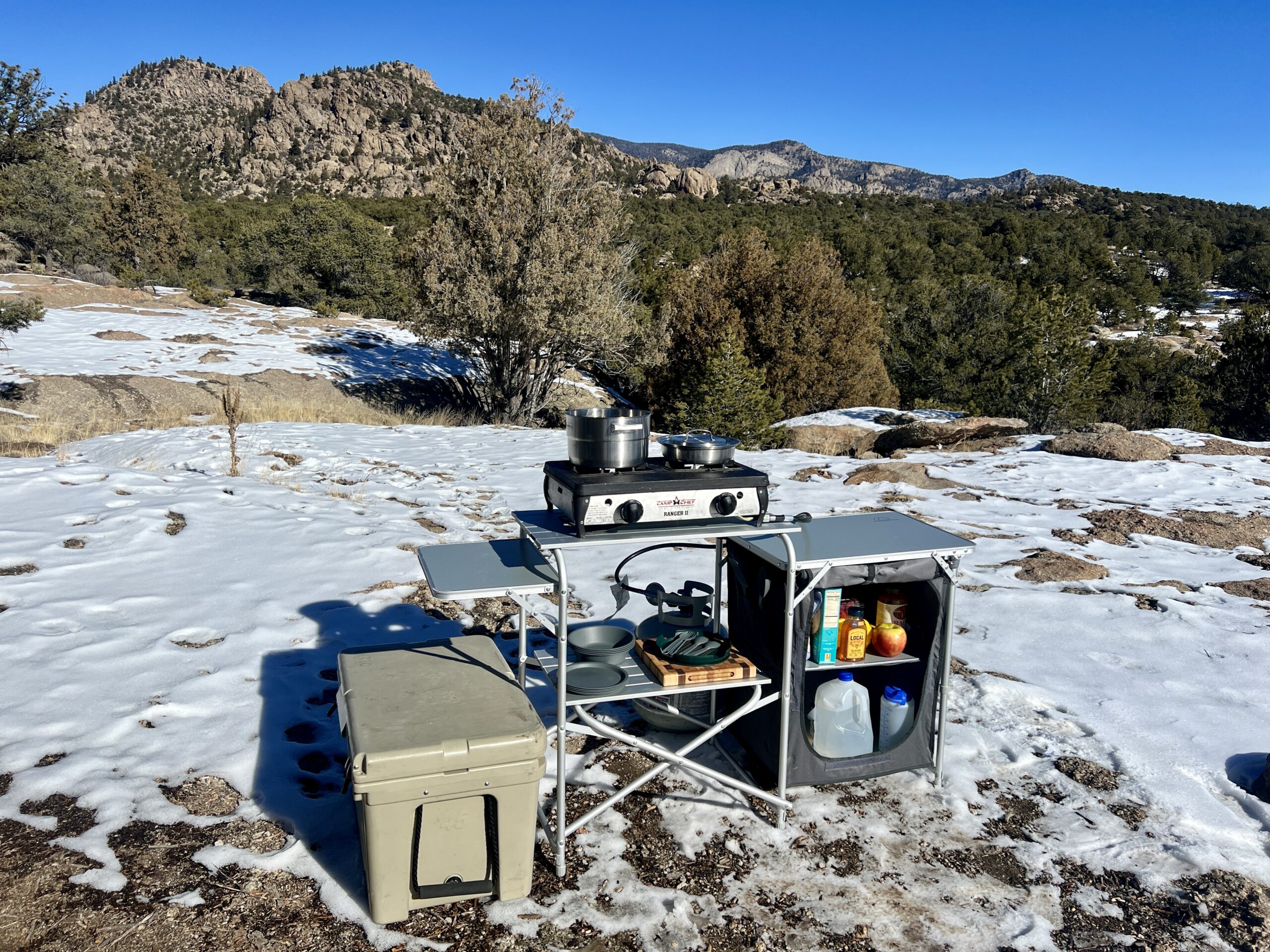 The Mountain Summit Gear Deluxe Roll Top Kitchen stands on snowy ground with rocky mountains and trees in the background. The table is loaded with a two-burner stove, cookware, and food items. A cooler is sitting next to it.