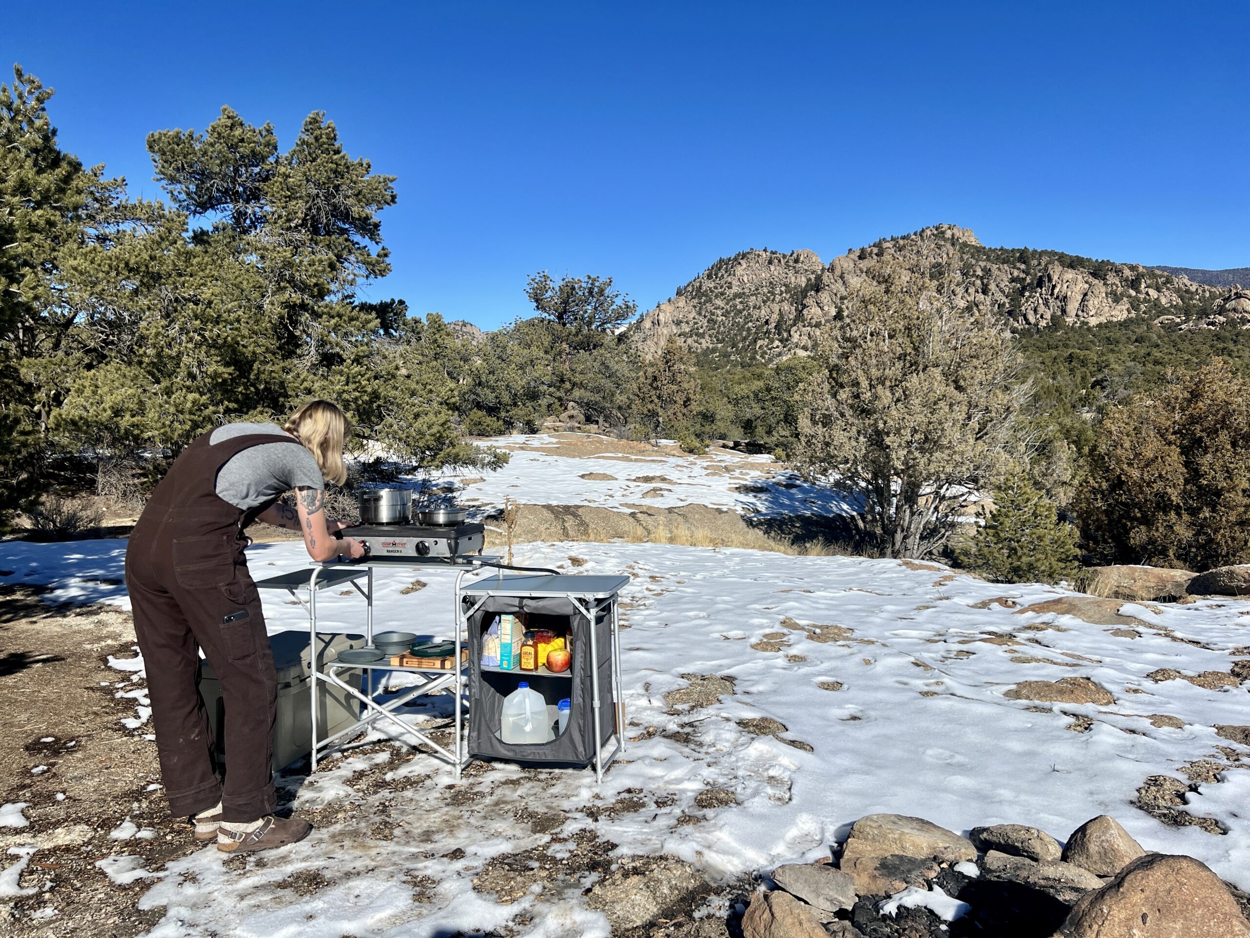 The Mountain Summit Gear Deluxe Roll Top Kitchen stands on snowy ground with a scenic backdrop of rocky mountains and oak trees. A person stands at the table touching a two-burner stove and the table has food items in its removable compartment.