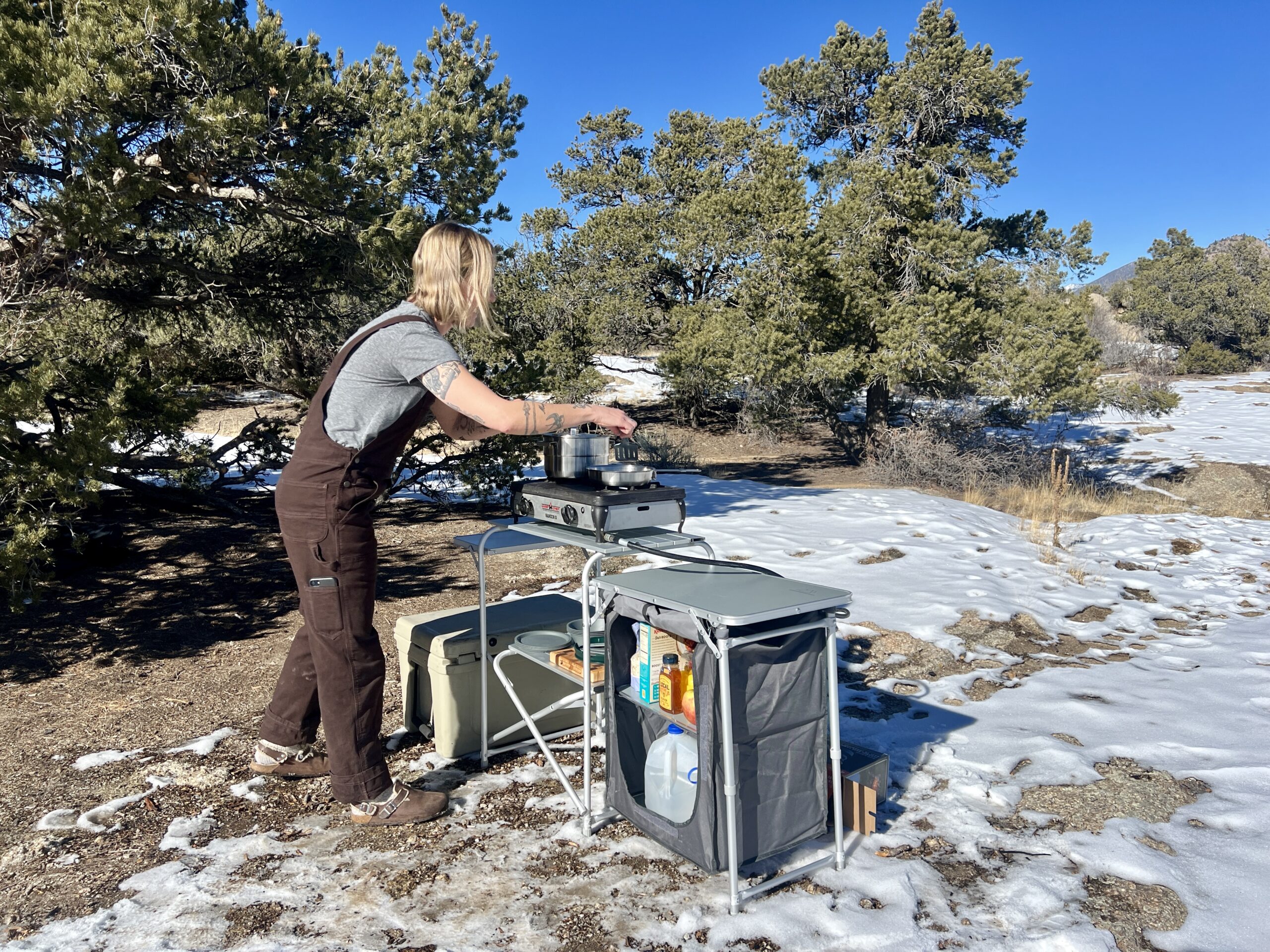 The Mountain Summit Gear Deluxe Roll Top Kitchen stands on snowy ground with oak trees behind it. A person in brown overalls and a gray t-shirt stands at the table cooking.