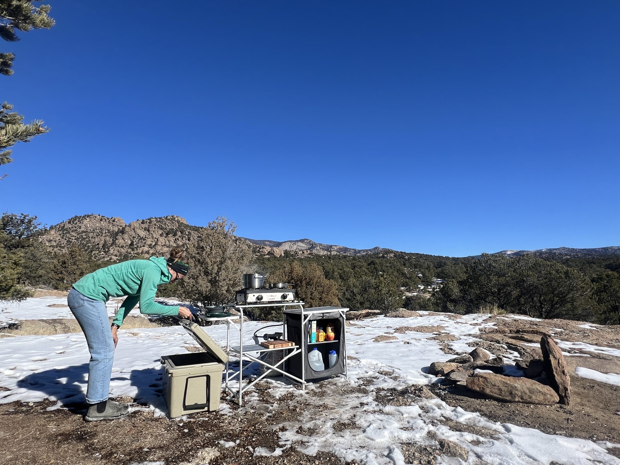 The Mountain Summit Gear Heavy Duty Roll Top Kitchen stands in snow and dirt with a backdrop of rocky hills and oak trees. A person standing next to the table wearing jeans, and a teal sweater, is looking into an ice chest.