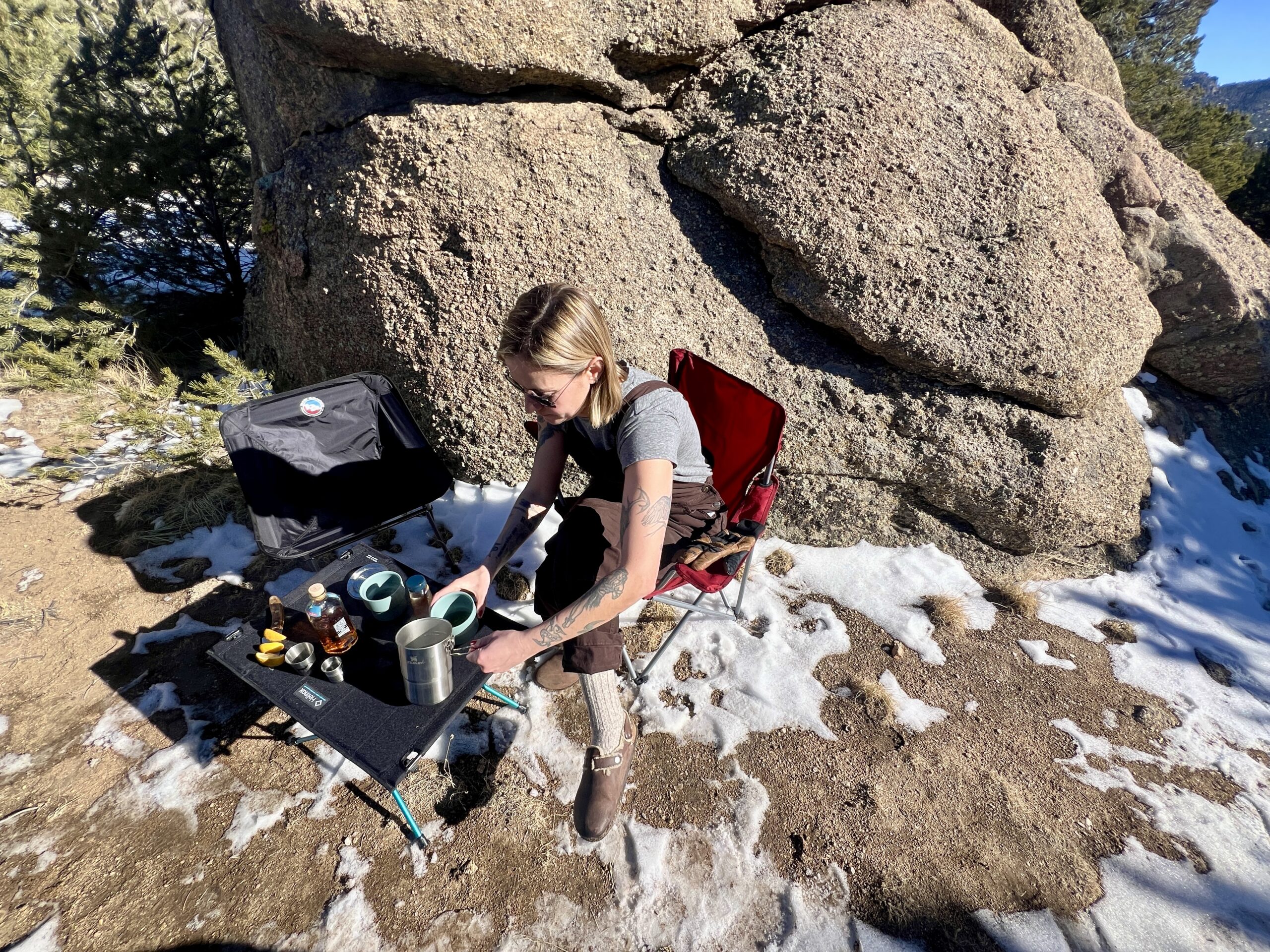A person sits in a red chair at the Helinox One Hard Top and pours a beverage into a cup. The table has whiskey and sliced lemons on top of it. An empty back chair is on the left. The background is a large boulder, trees, and snow.