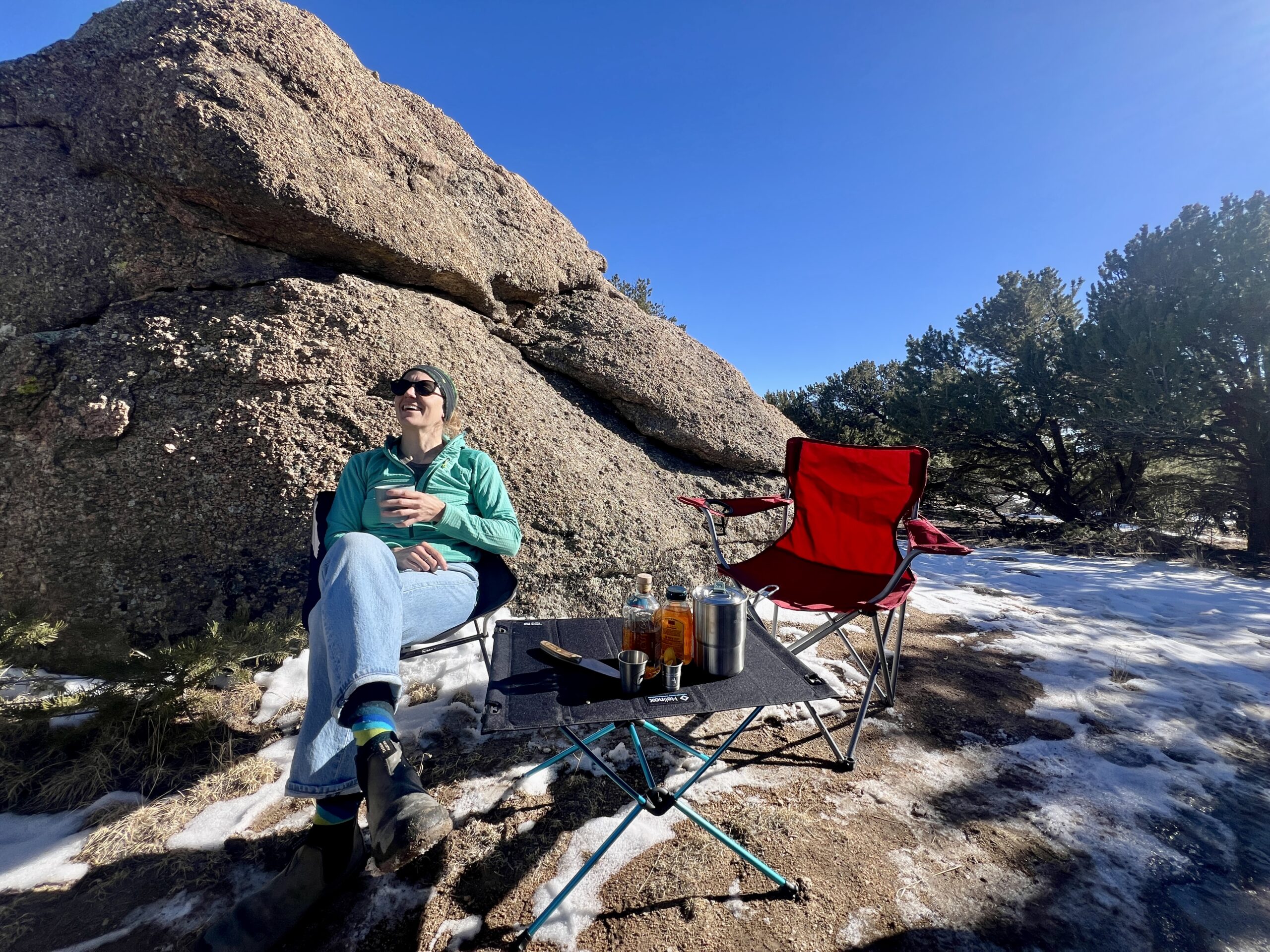 The Helinox Table One Hard Top has beverages on it. A person is smiling and looking ahead with a teal sweater, sunglasses, and jeans on. The backdrop has trees and a large boulder and there is snow on the ground. A red chair is on the right.