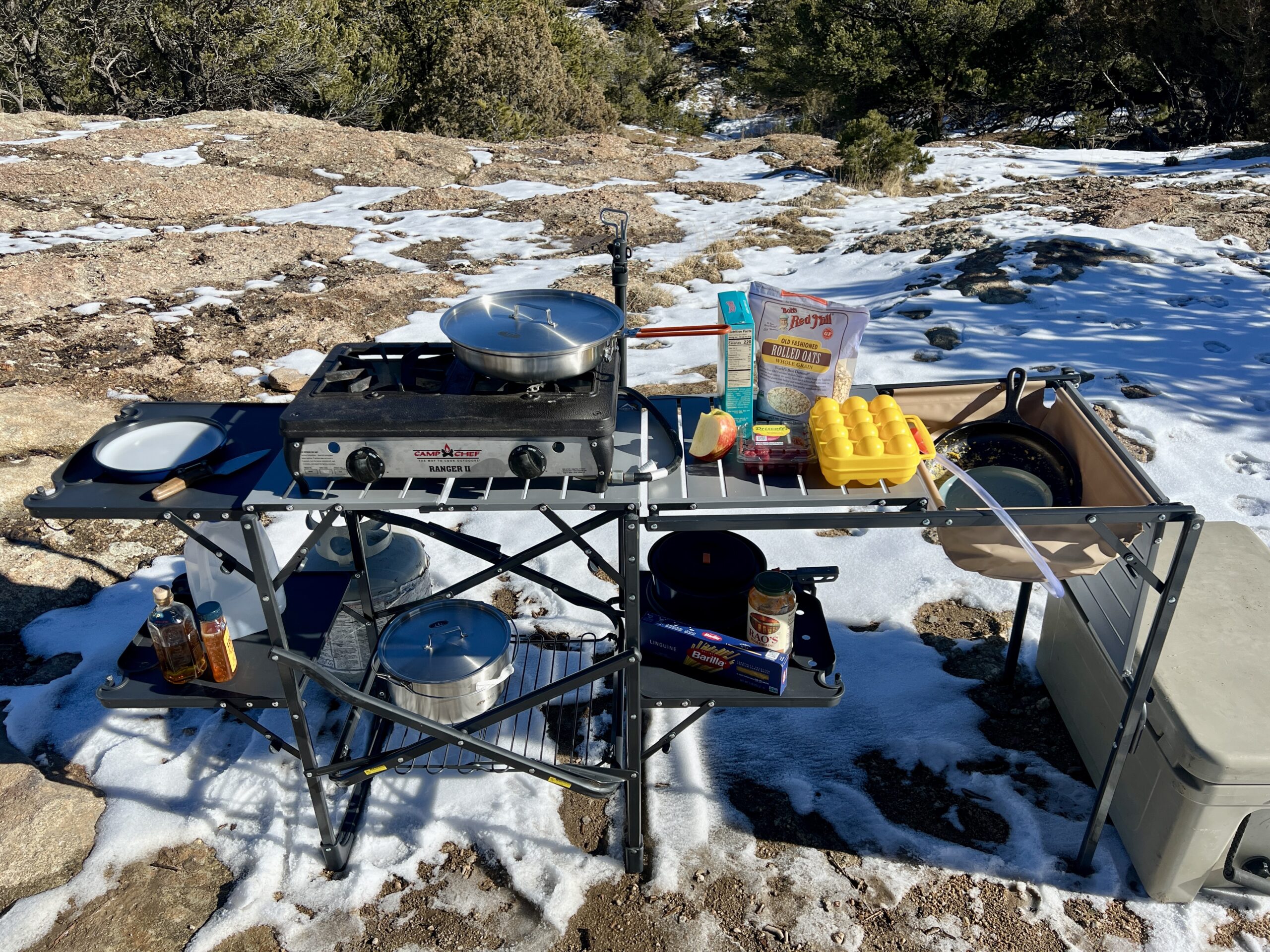 The GCI Master Cook Station stands outside in snow and rocky terrain. Its shelves are full of kitchen items with a sink full of dirty dishes. A cooler is on the ground next to it.