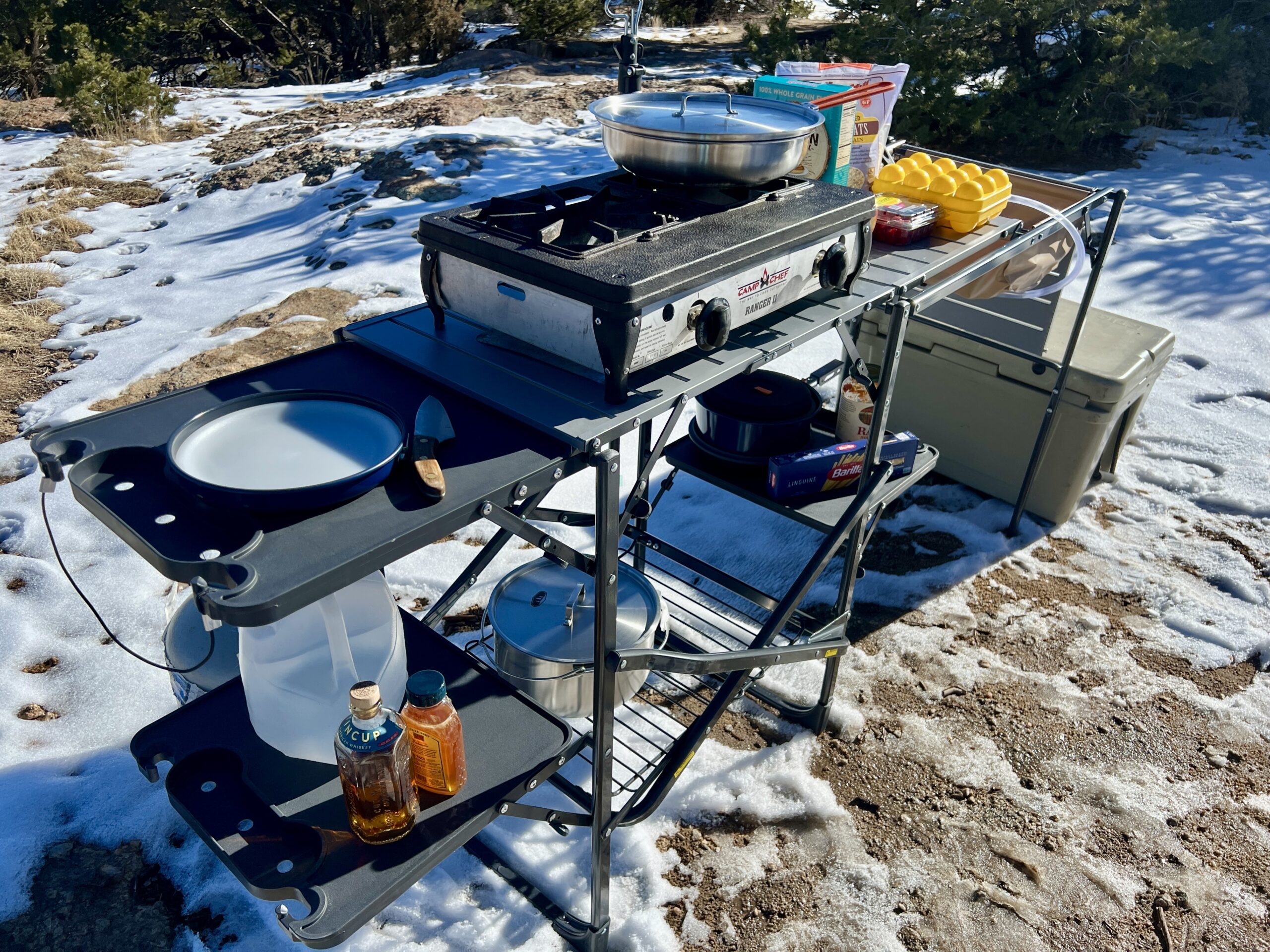 The GCI Master Cook Station stands outside in snow and rocky terrain. Its shelves are full of kitchen items. A cooler is on the ground next to it.