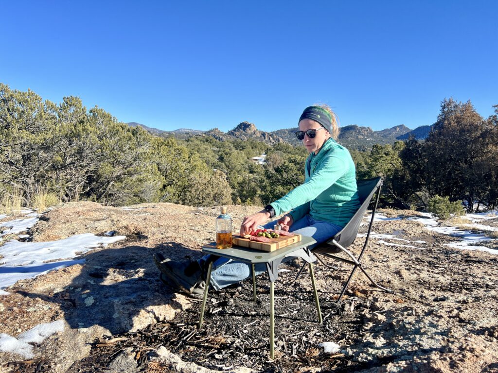 A person sits in a camp chair in a mountain setting next to the NEMO Moonlander Dual-Height Camp Table. She is reaching for food on a charcuterie board that is on the table.