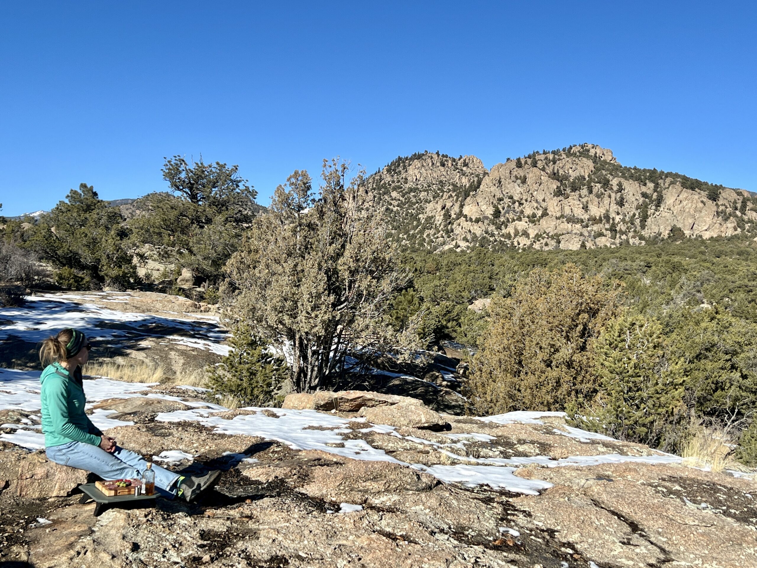 A person sits on a rock looking out into the mountains. Next to her, the NEMO Moonlander Dual-Height Camp Table sits on a rock, with a charcuterie board on top of it.