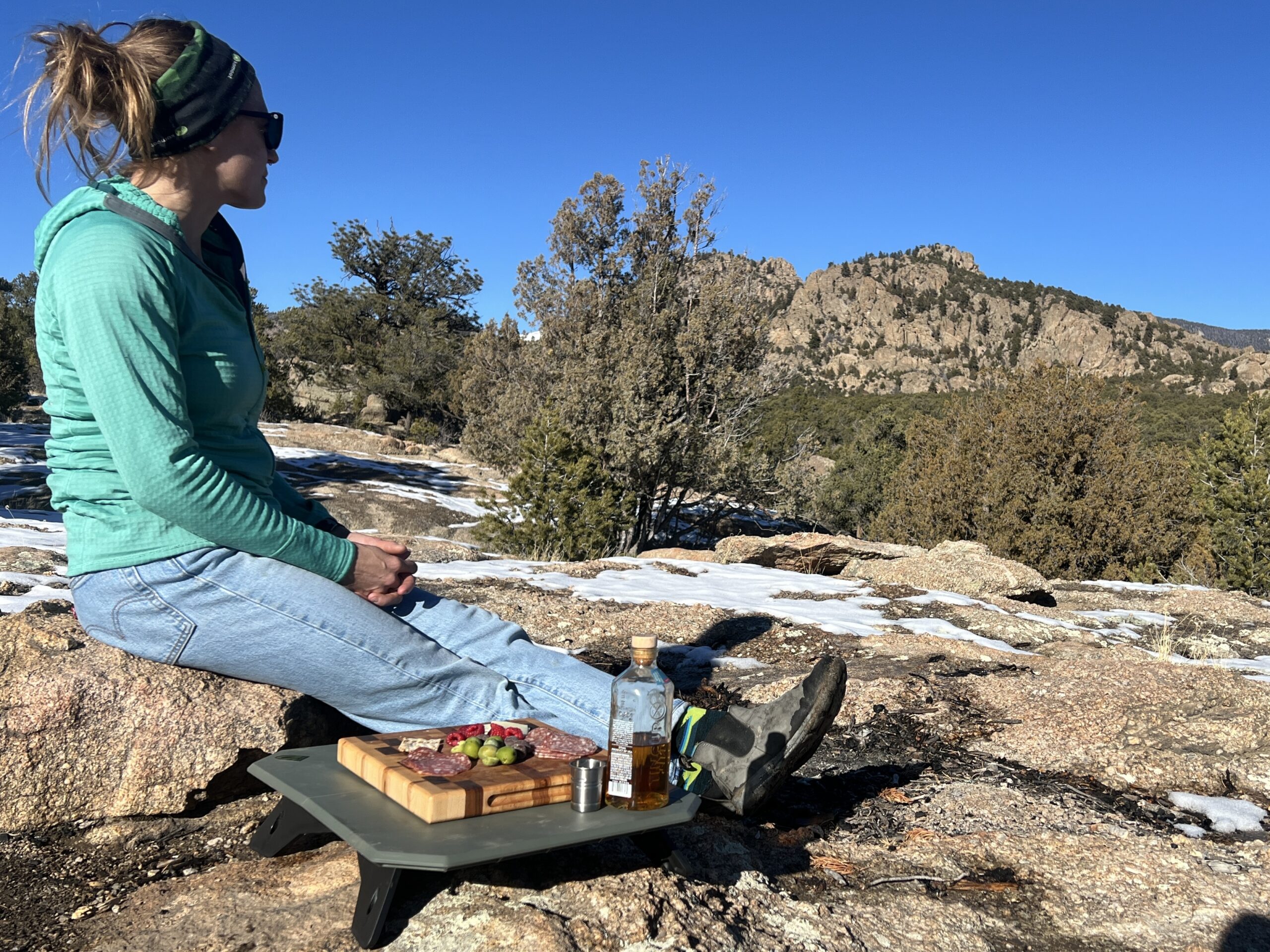 A person sitting on a rock looking out into the distance. The NEMO Moonlander Dual-Height Camp Table is sitting on a rock next to her with a bottle of whiskey and a charcuterie board on top of the table.