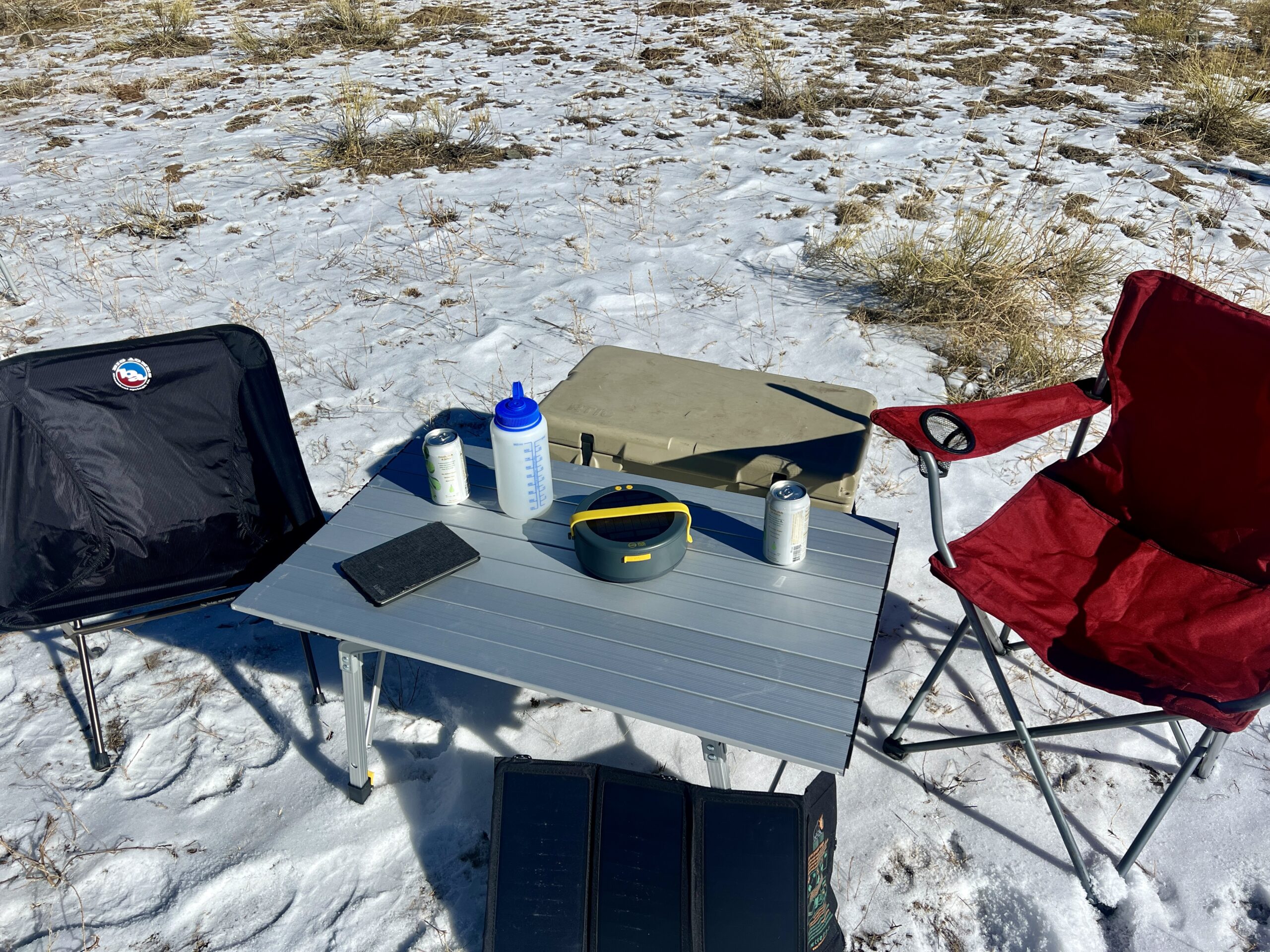The Portal Quick Adjust Lightweight Roll-Up Table is surrounded by a red chair, black chair, and solar charger. On the top are a Nalgene, kindle, and cans. There is a beige cooler behind the table.