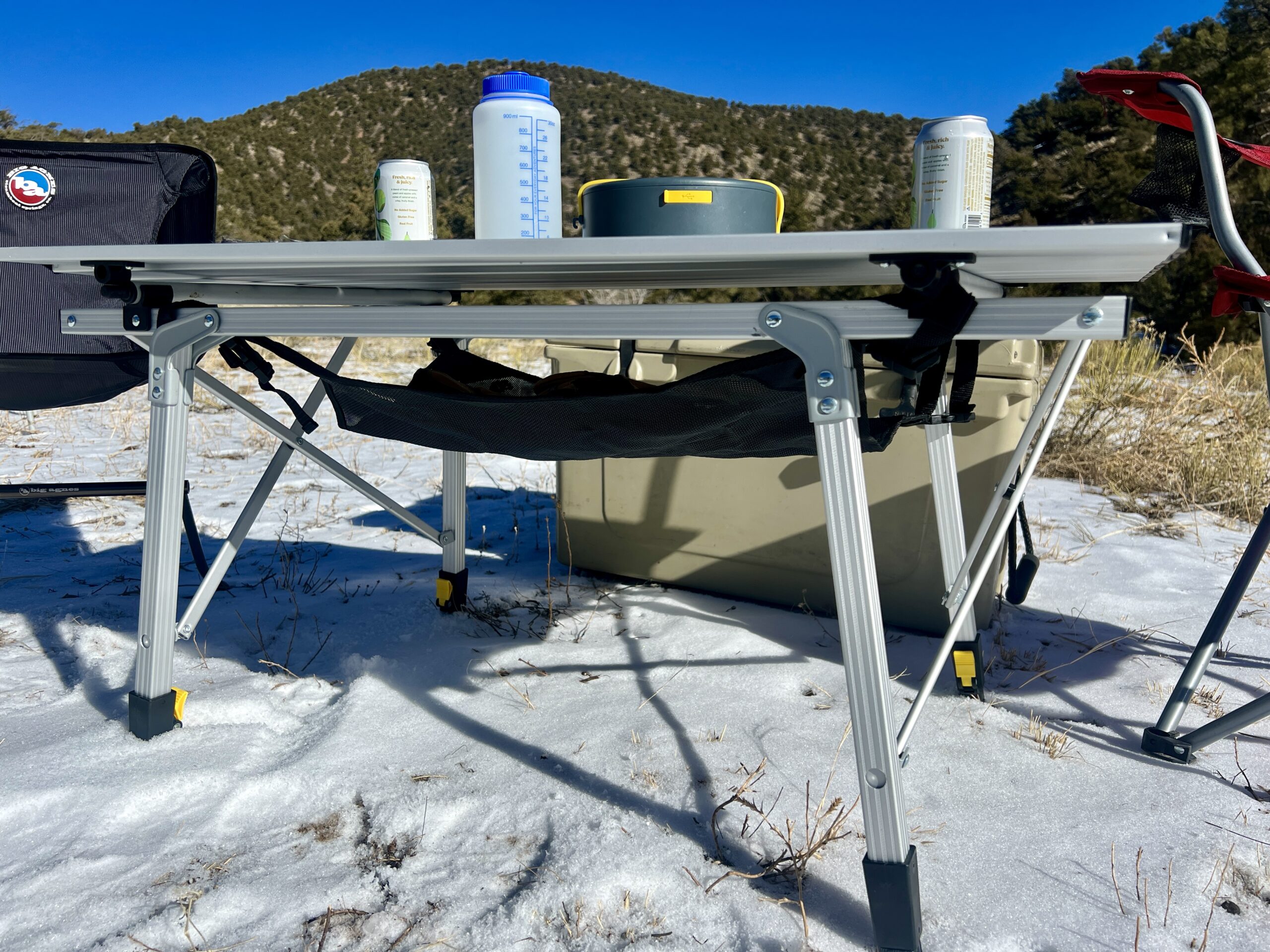 A close-up of the Portal Quick Adjust Lightweight Roll-Up Table. On top of the table is a Nalgene, solar charger, and cans. A beige cooler is behind the table. The background is tree-covered hills.