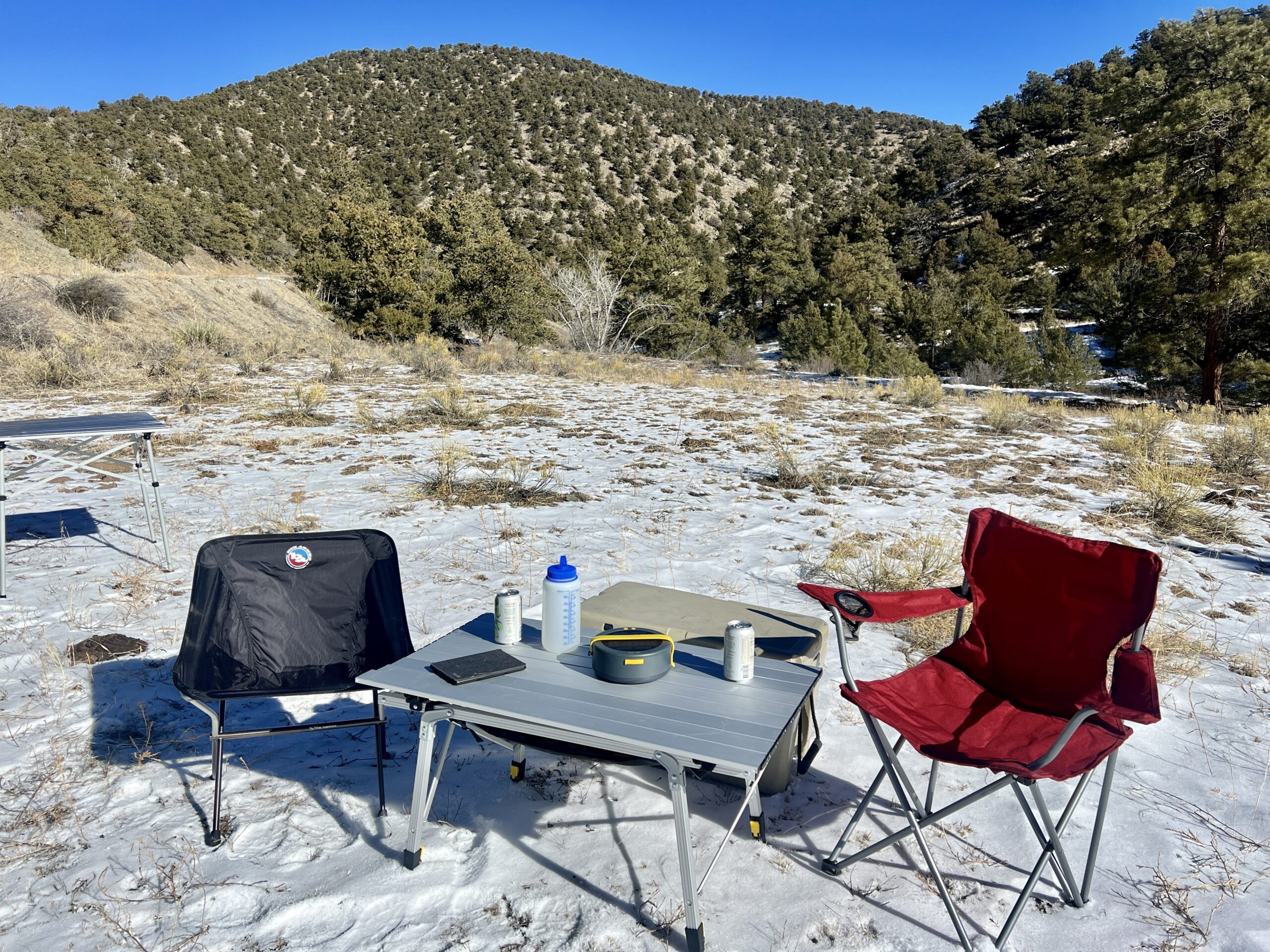The Portal Quick Adjust Lightweight Roll-Up Table is surrounded by a red chair, black chair, and solar charger. On the top are a Nalgene, kindle and cans. The background