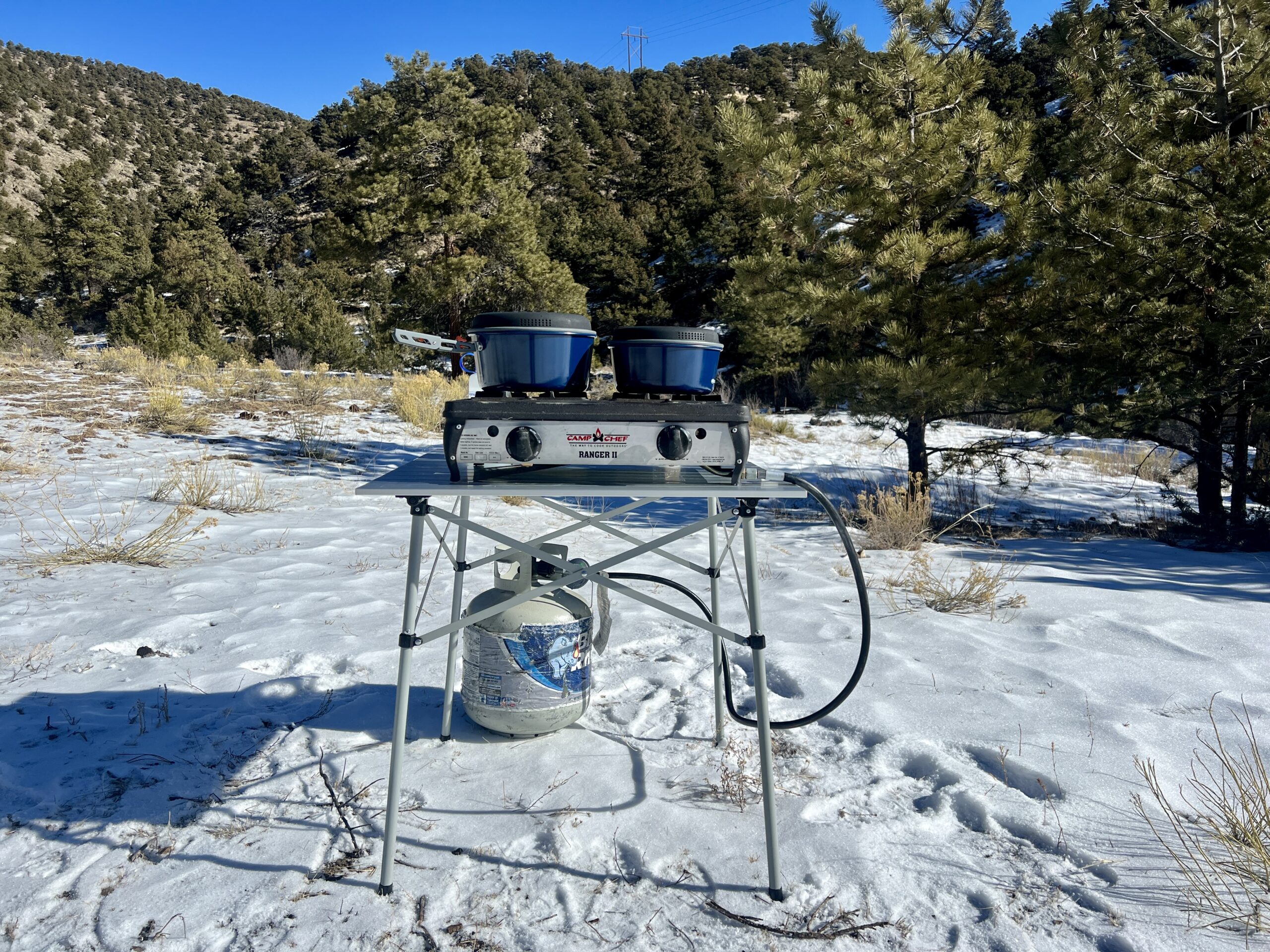 The Coleman Compact Roll-Top Table stands on snow with a two-burner stove and blue cookware on its top. A propane tank sits on the ground behind the table. The background is oak-tree covered hills.