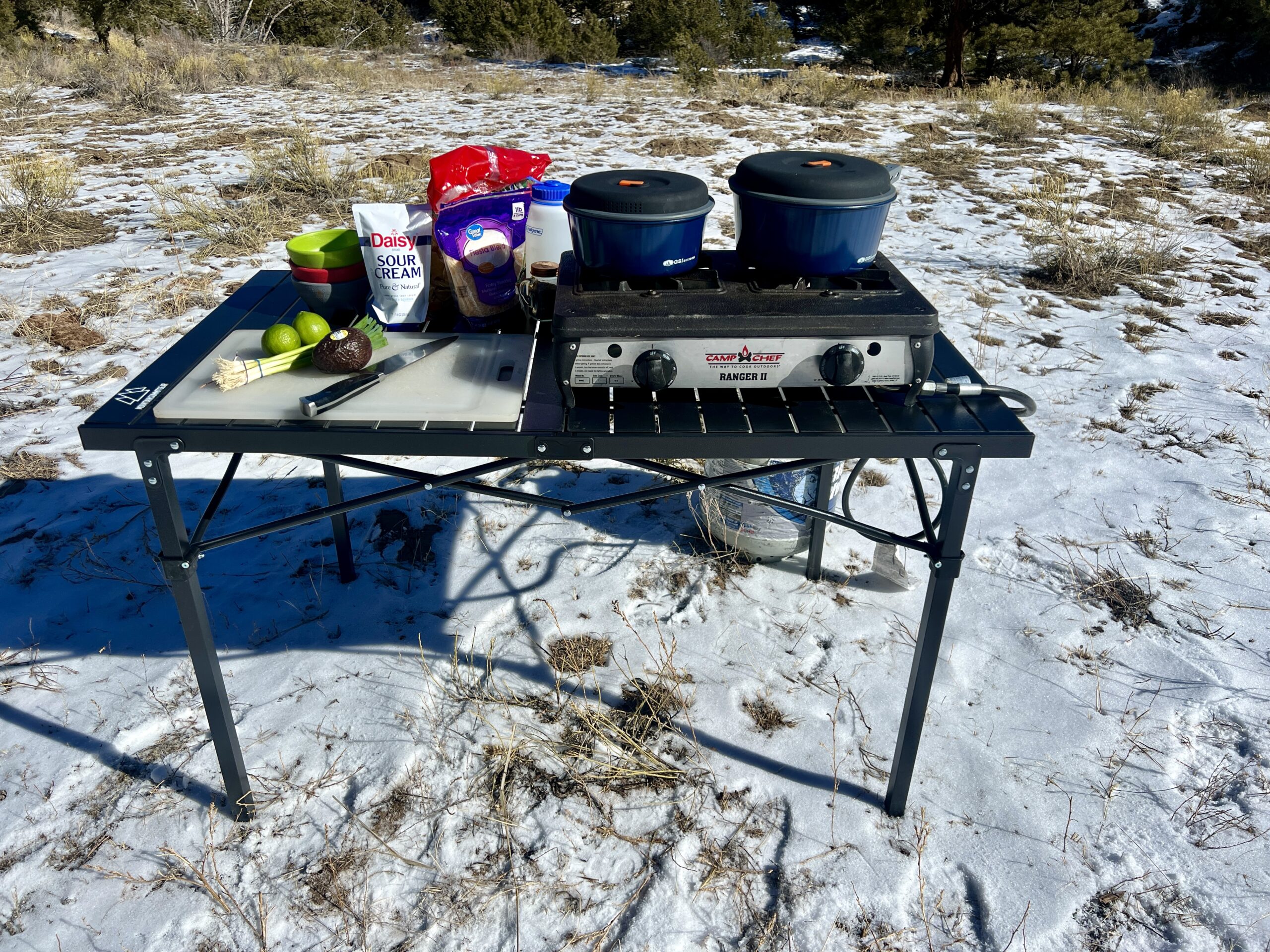 The Mountain Summit Gear Heavy Duty Roll Top Table stands in the snow with tree-covered hills in the background. A two-burner stove and food items are on top. A propane tank sits on the ground behind it.