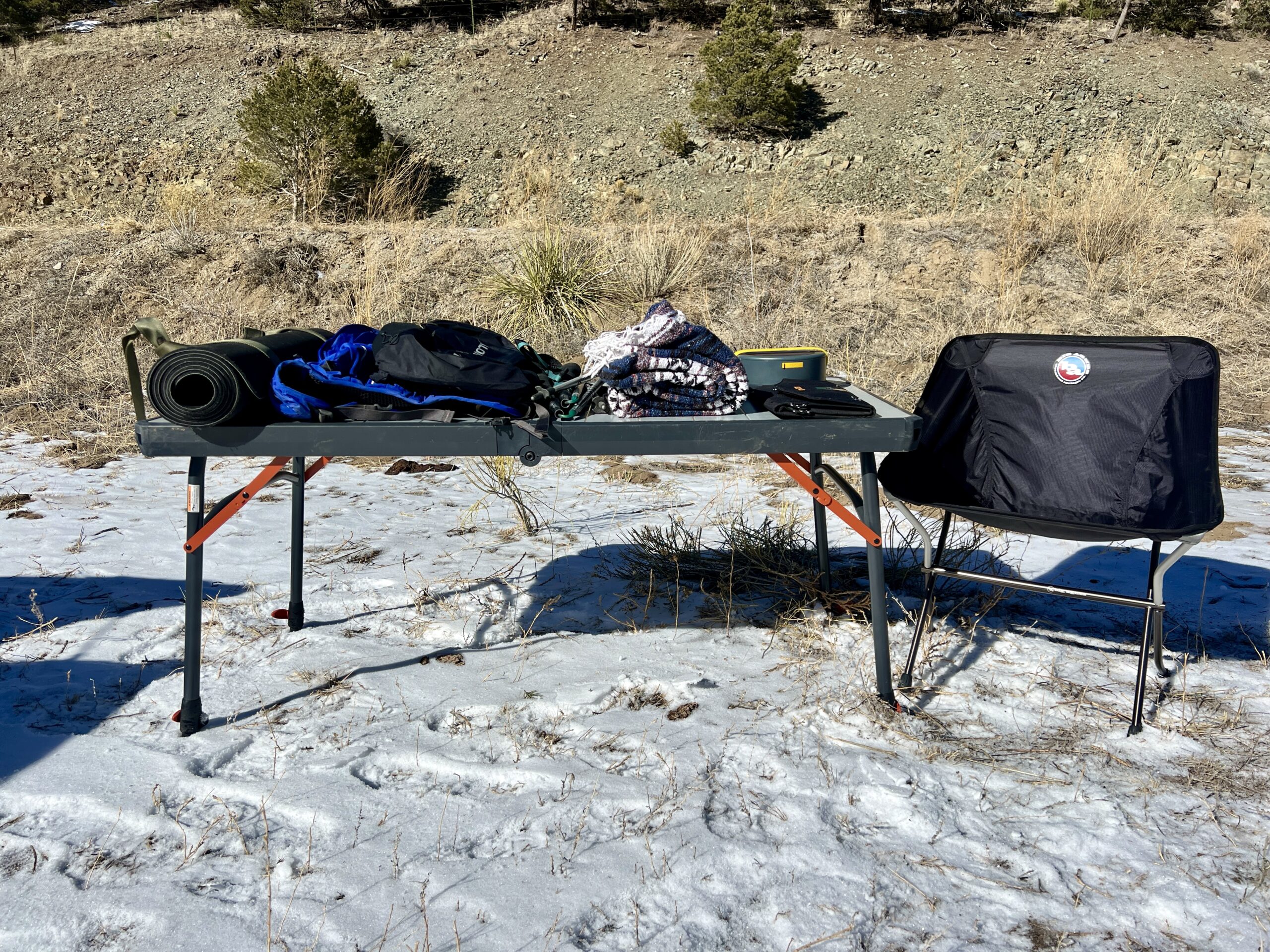 The REI Camp Prep Table stands in the snow with a yoga mat, blue and black backpacks, hiking poles, a blanket, and solar chargers. A black camp chair is on the right of the table.