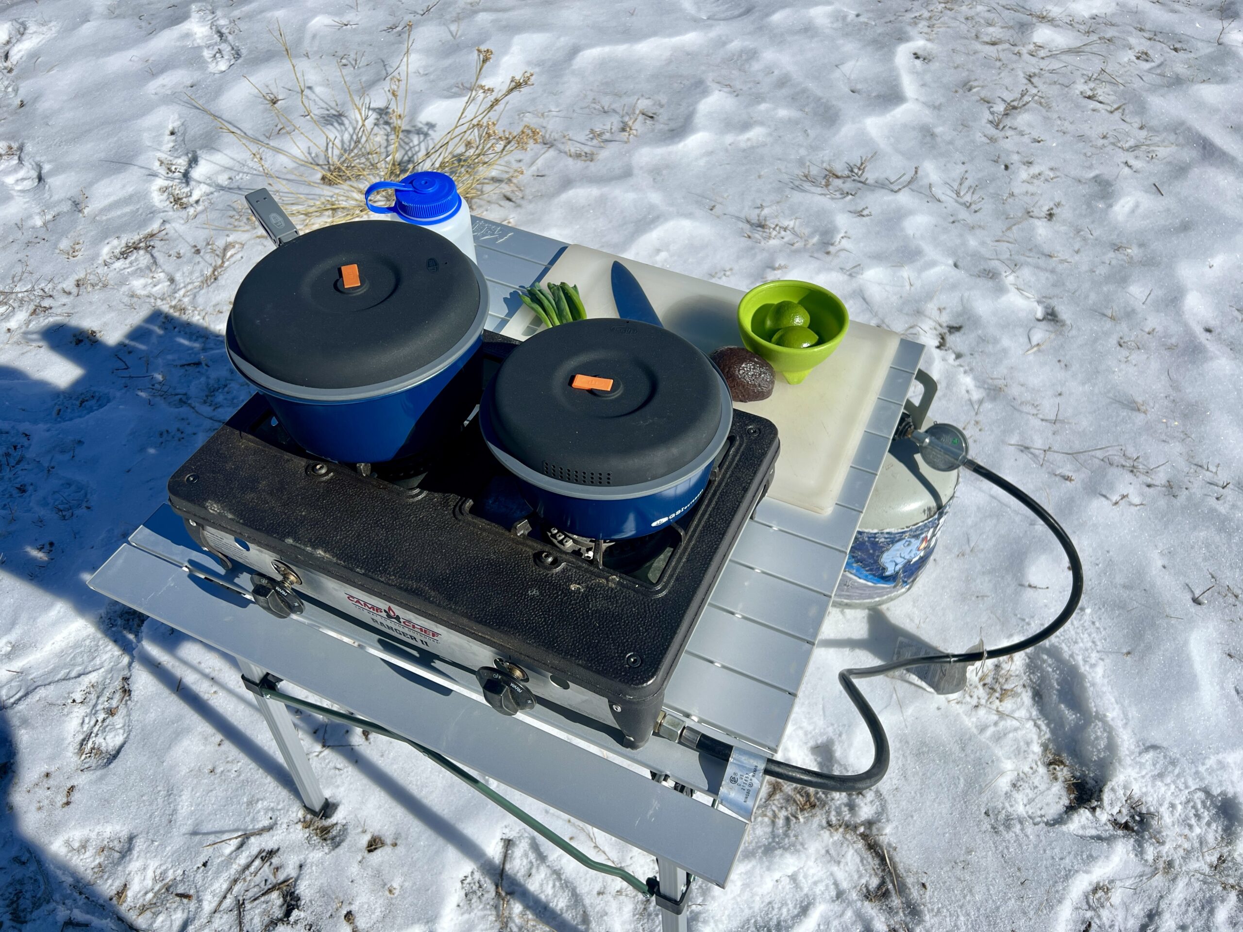 The REI Camp Roll Table stands in the snow and holds a two-burner stove, cookware, a cutting board, a knife, an avocado, scallions, a Nalgene, and a bowl with limes on top.