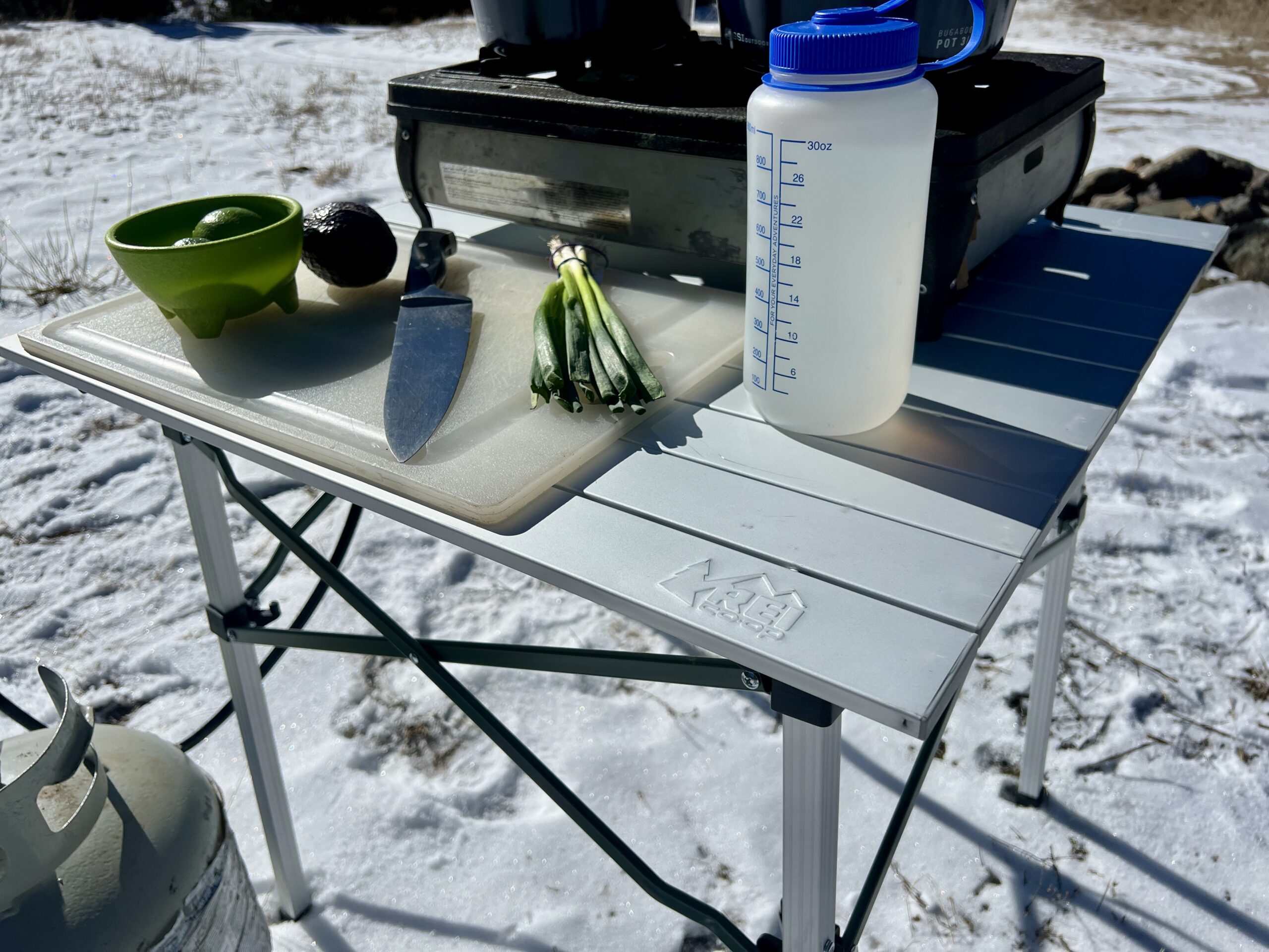 The REI Camp Roll Table stands in the snow. It has a Nalgene, cutting board, knife, scallions, green bowl, limes, and two-burner stove on top.