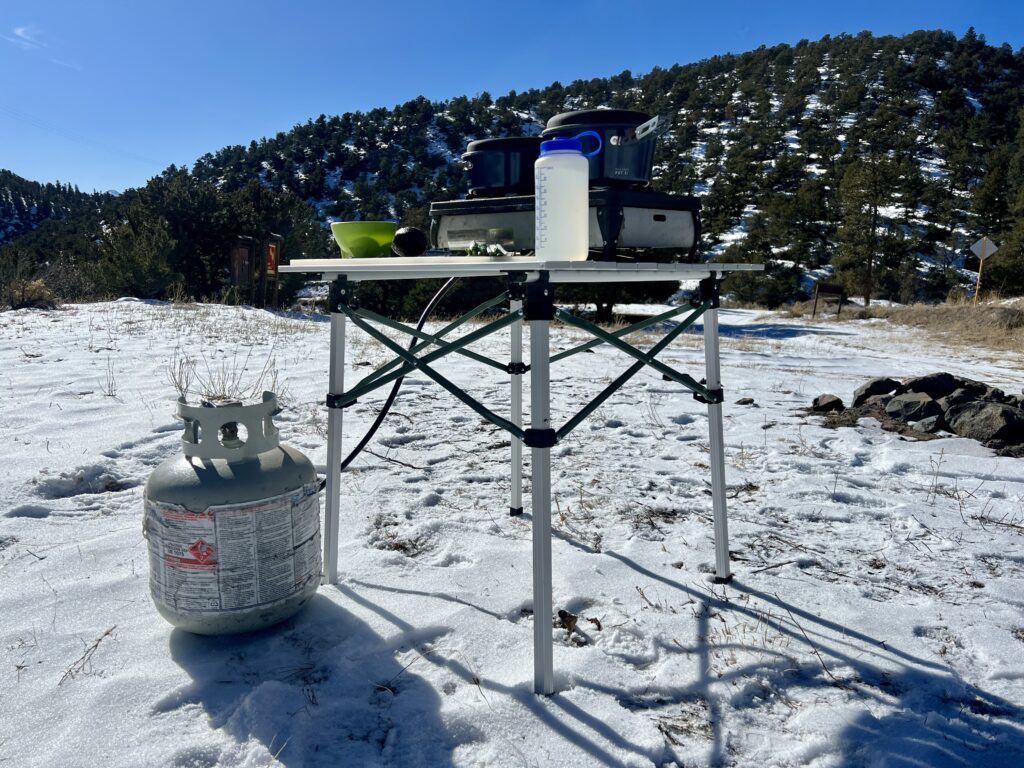 The REI Camp Roll Table stands in the snow with a tree-covered hill in the background. It has a Nalgene, two-burner stove, green bowl, and an avocado on its top. A propane stove sits on the ground next to the table.