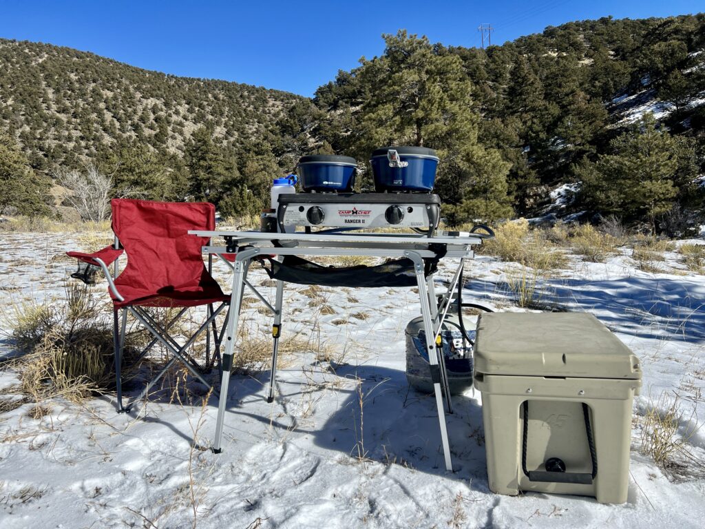 The Portal Roll-Up Table is standing on snow with a stove and cookware on top of it. There is a beige cooler and a red chair next to it. The background is tree-covered hills.