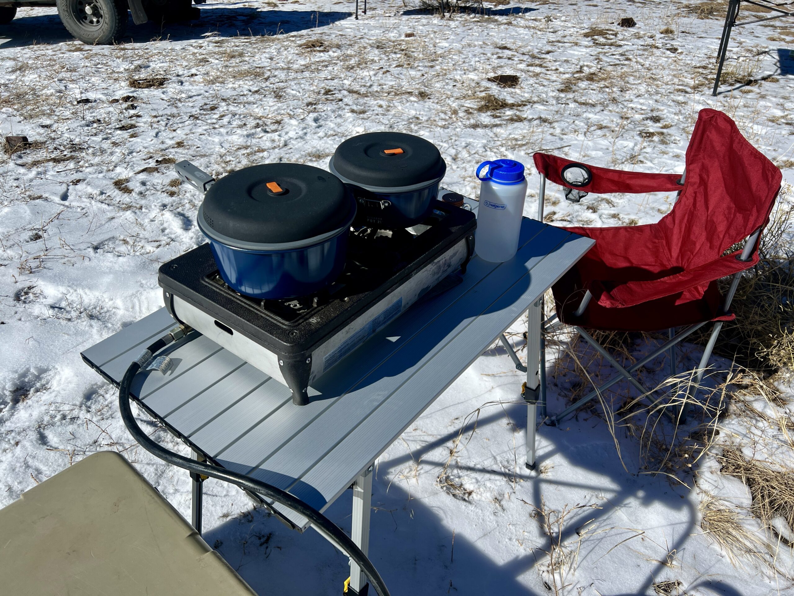 The Portal Roll-Up Table is standing in the snow with a two-burner stove and blue cookware on top of it. A red chair sits next to the table.
