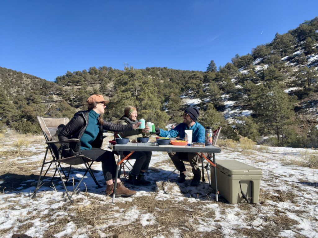 Three people are cheers-ing and smiling at each other. The REI Camp Prep Table is in the center with bowls and food on top of it. There's a beige cooler on the right. The background is hills covered in trees and snow.