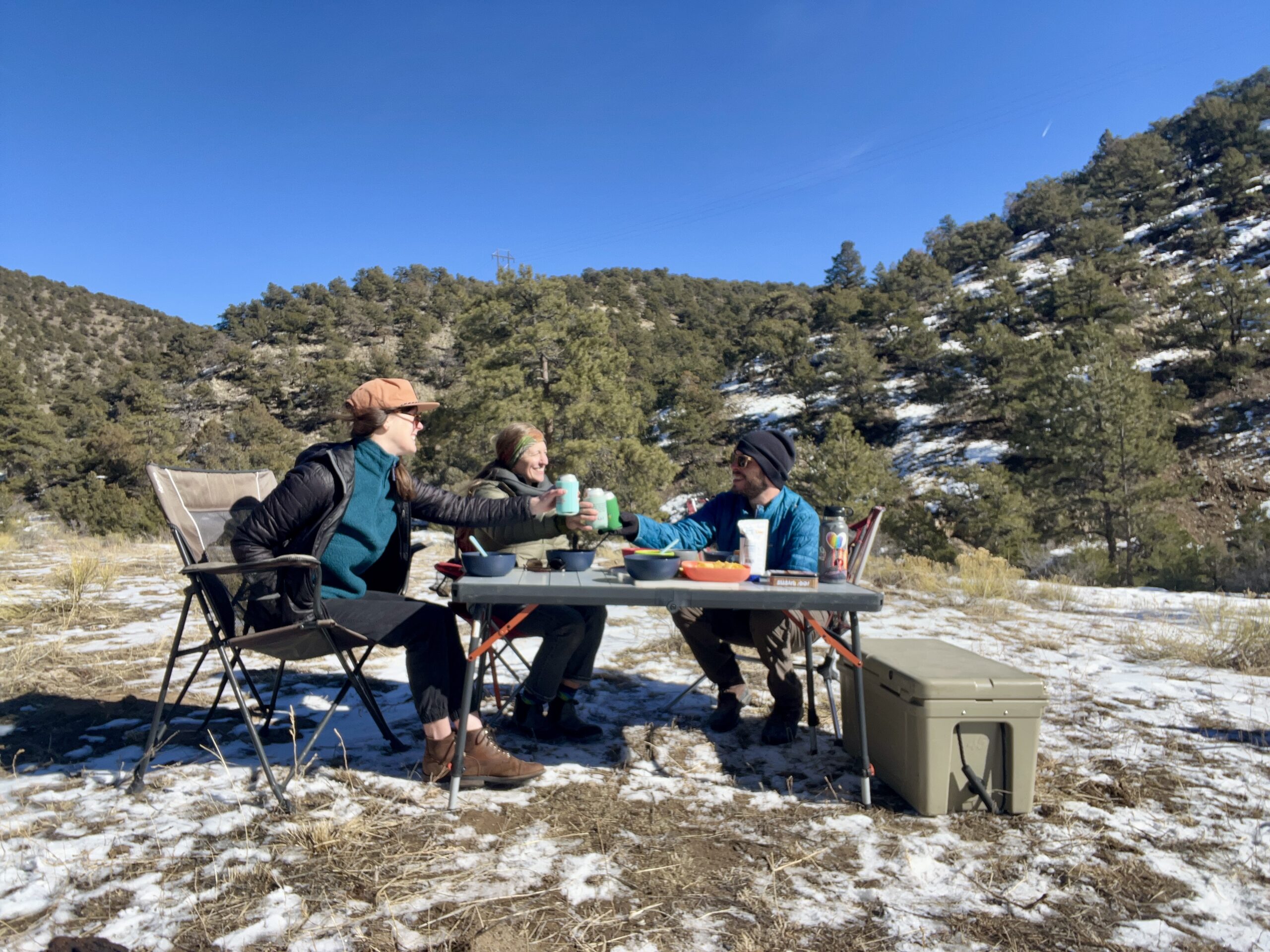 Three people are cheers-ing and smiling at each other. The REI Camp Prep Table is in the center with bowls and food on top of it. There's a beige cooler on the right. The background is hills covered in trees and snow.