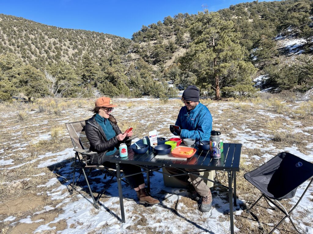 Two people sit and smile around the Mountain Summit Gear Heavy Duty Roll Top Table. They are wearing black and blue jackets and black and orange hats. They have playing cards in their hands. There are bowls, food, and drinks on the table.