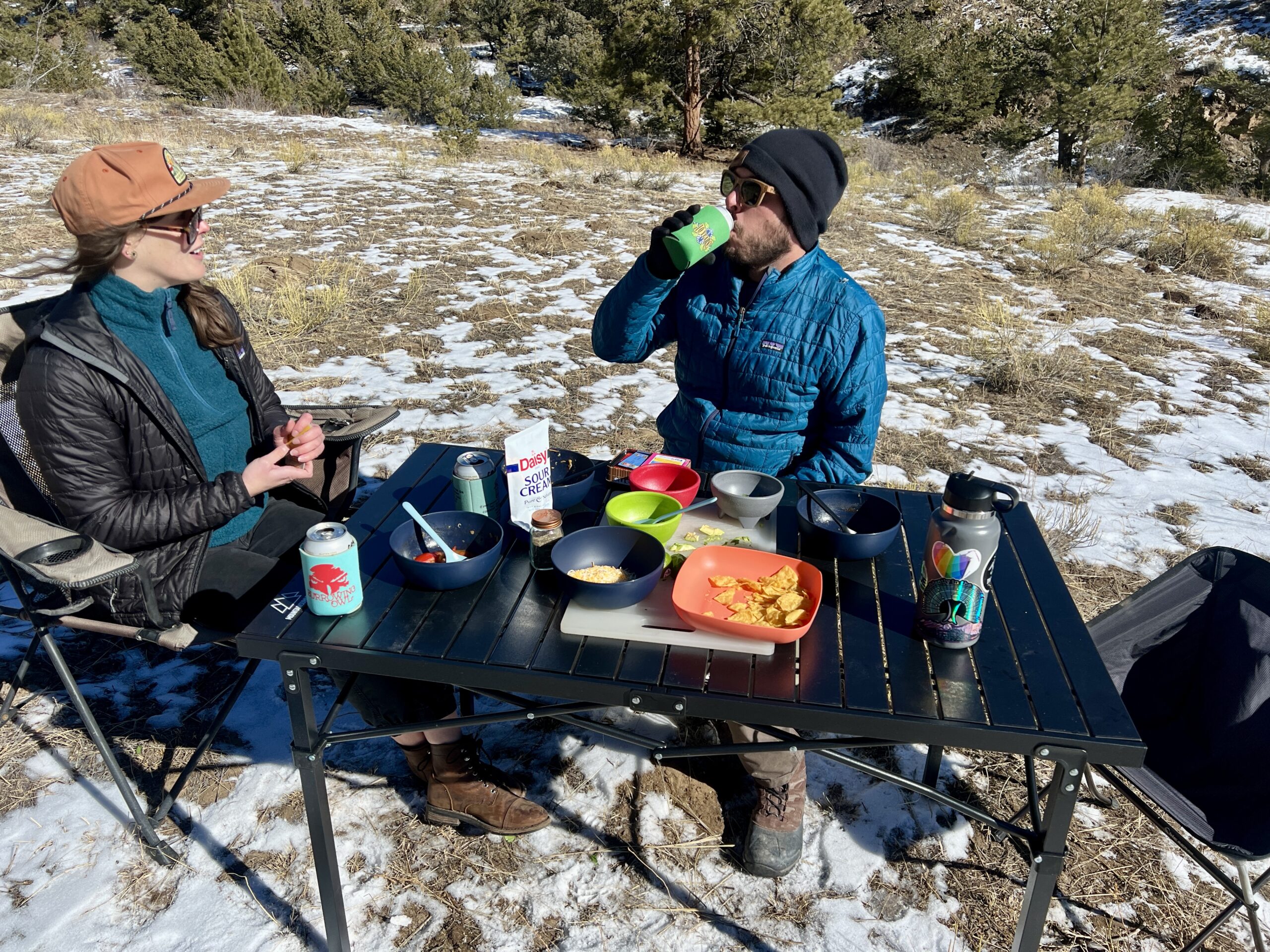 Two people sit and smile around the Mountain Summit Gear Heavy Duty Roll Top Table. They are wearing black and blue jackets and black and orange hats. They have playing cards in their hands. There are bowls, food, and drinks on the table.