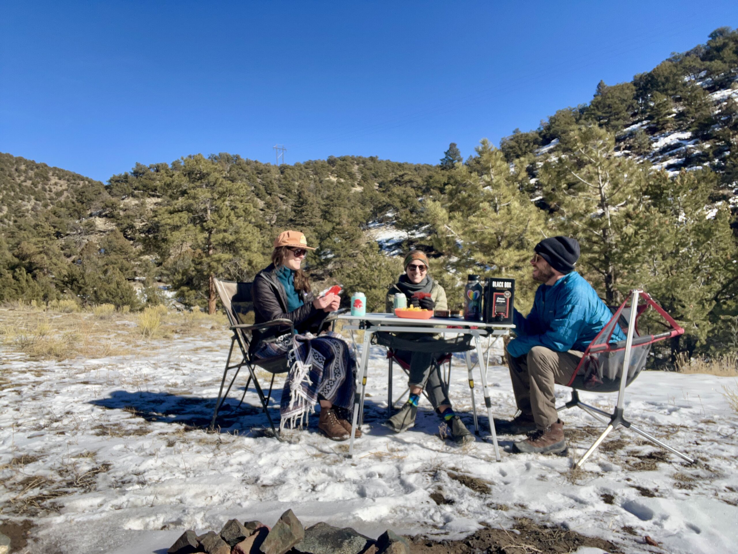 Three people sit around the Portal Roll-Up Table smiling and holding cards. The background is tree-covered hills and there is snow on the ground.