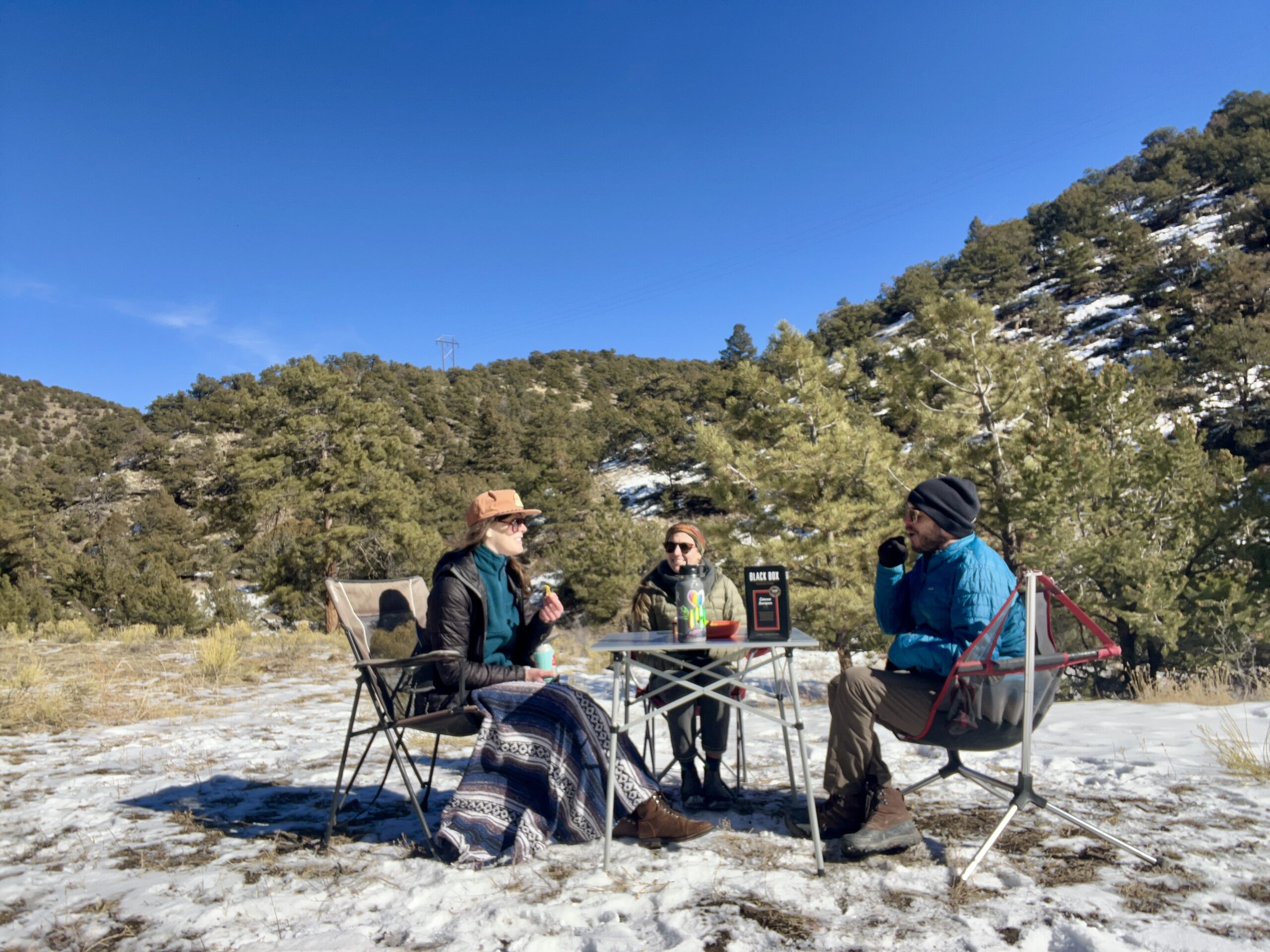 Three people sit around the Coleman Compact Roll-Top Table and are smiling. The background are snow and oak-covered hills. The table has a box of wine and a water bottle.