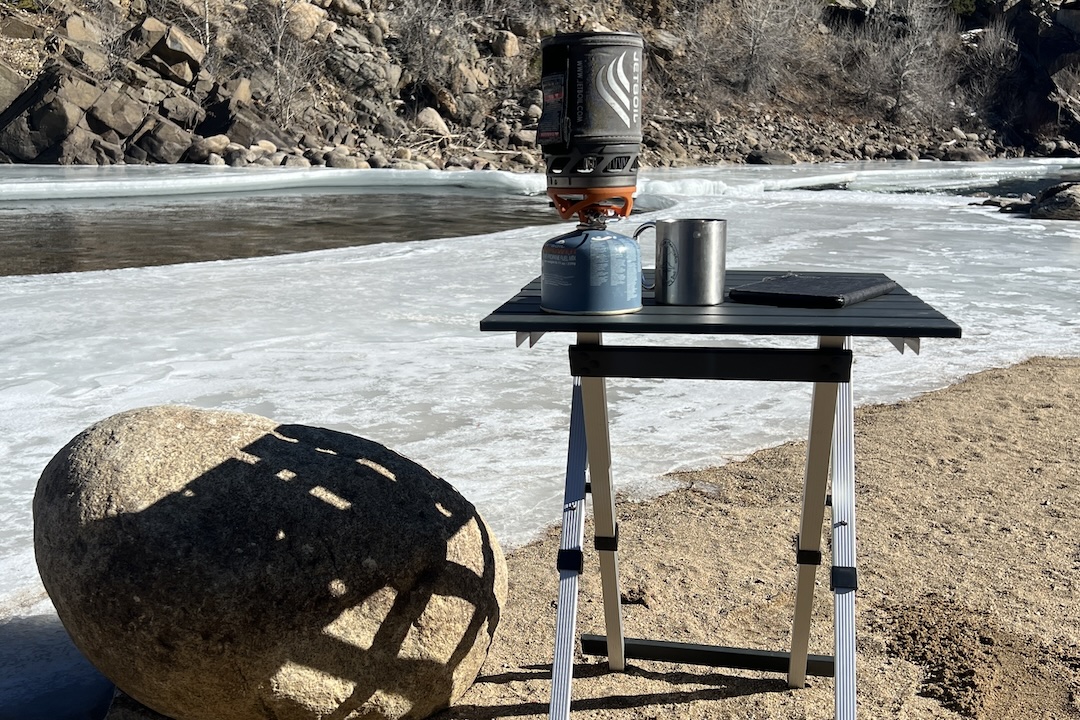 The GCI Compact Table 20 stands in the sand along a river. Ice is covering most of the river. A large boulder is next to the table.