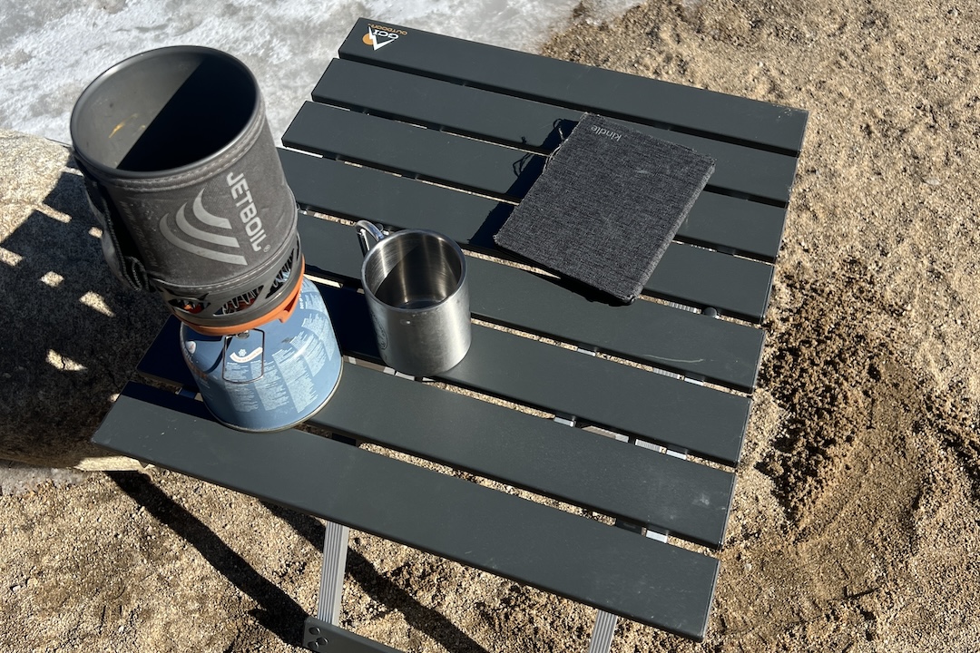 The GCI Compact Table 20 stands on the sand. A jetboil, stainless cup, and kindle sit on top.