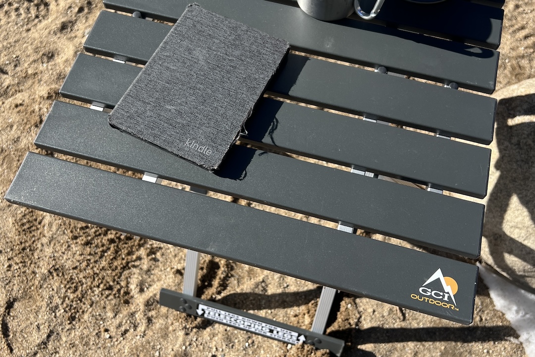 The GCI Compact Table 20 stands in the sand. It has a visible Kindle and stainless cup on it.