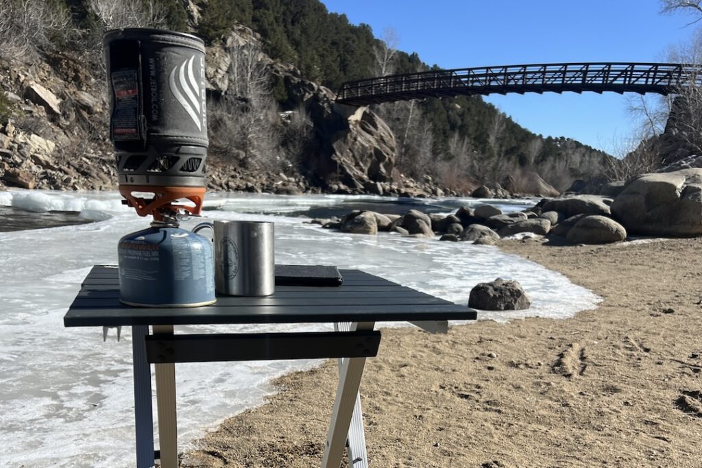 The GCI Compact 20 stands on a sandy river beach. The river is mostly iced over. A Jetboil, stainless cup, and kindle sit on top of the table. In the background, a bridge crosses the river.