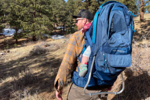 A man hiking through a forested area with the Kelty PerfectFIT Journey Signature carrier, showcasing its ergonomic design and sturdy kickstand.