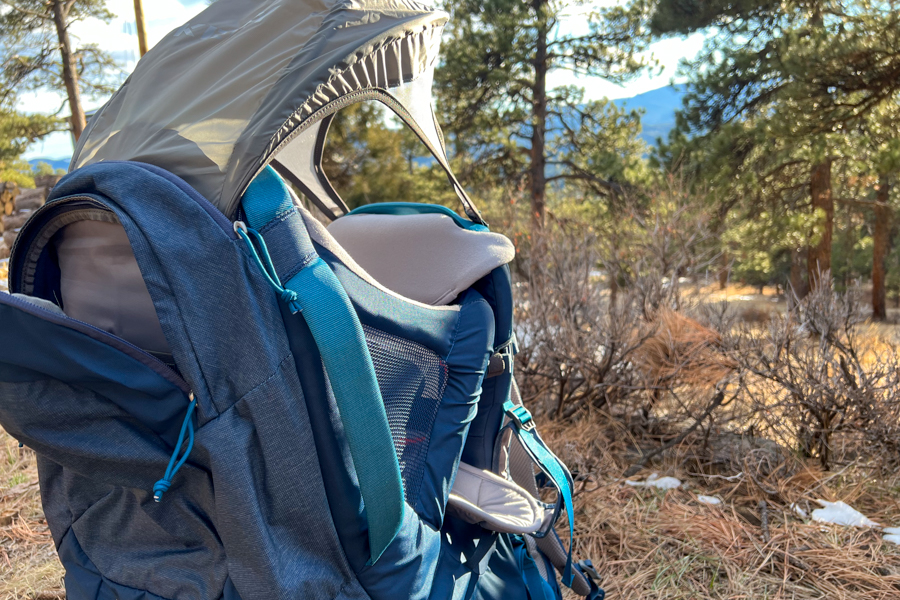 Side view of a baby carrier with an extended sunshade, set against a scenic forest background.