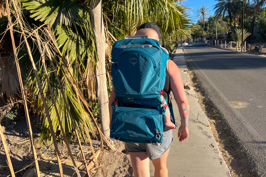 Back view of the Osprey Poco SLT child carrier, showing the storage compartments and sleek design as the wearer walks down a tropical road.