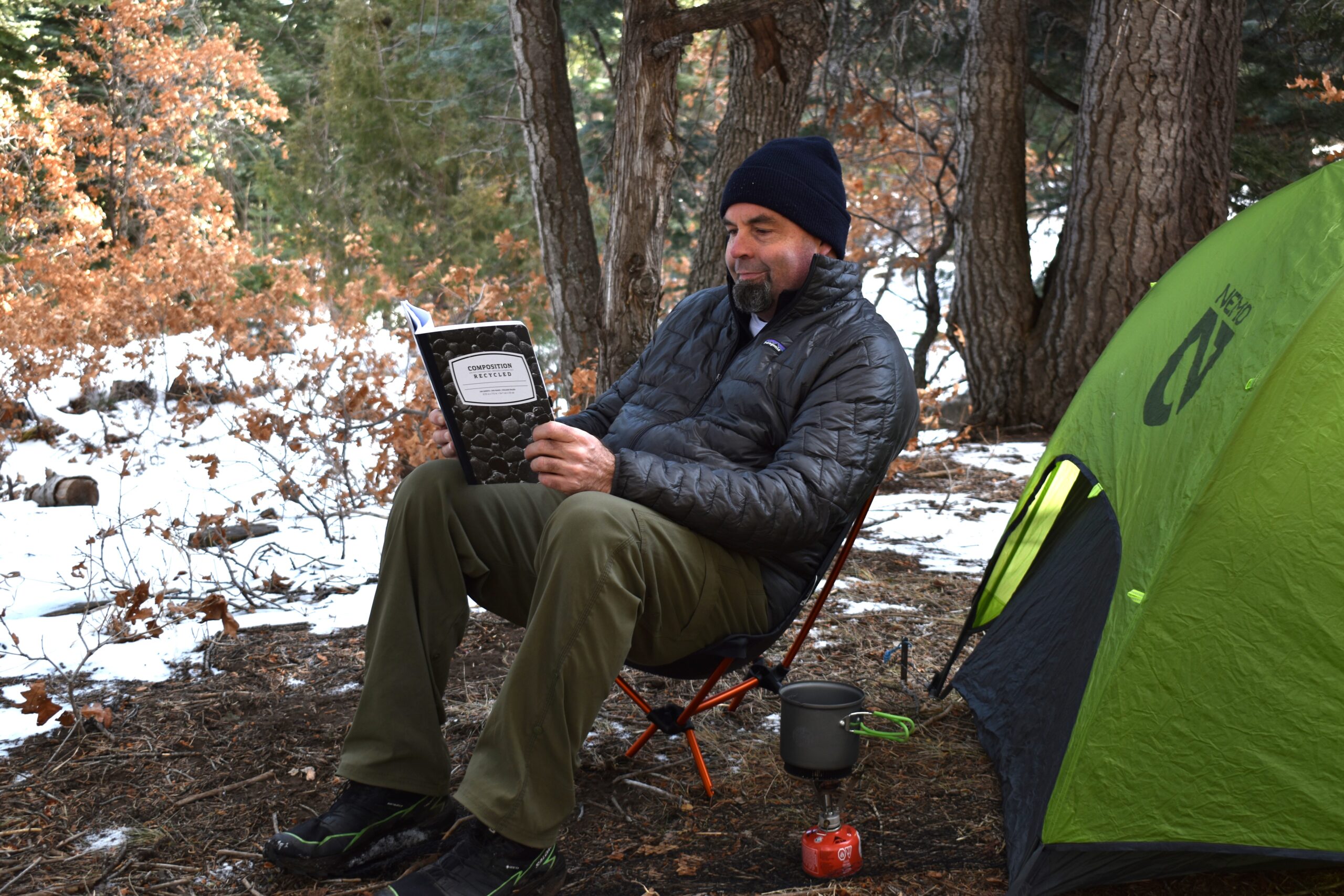A male backpacker reads a hiking journal while sitting on a backpacking chair next to a tent in the snow