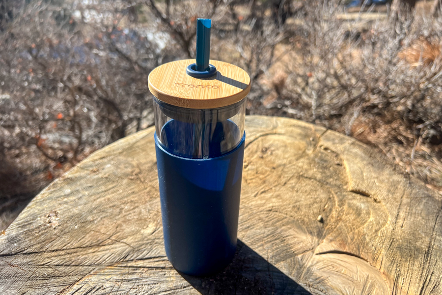 Tronco tumbler standing upright on a tree stump, showing its glass and bamboo design with a teal straw.