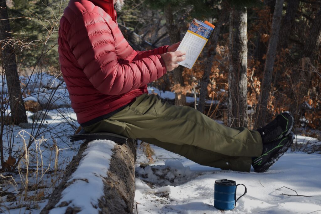 Hiker reads a guide book while sitting on a foam pad set on top of a log in the snow