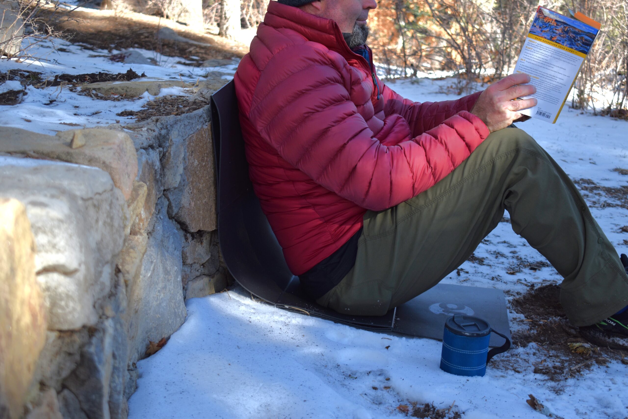a backer reads a hiking book while sitting on a pad as it keeps his butt off the snow and his back cushioned against a rock wall