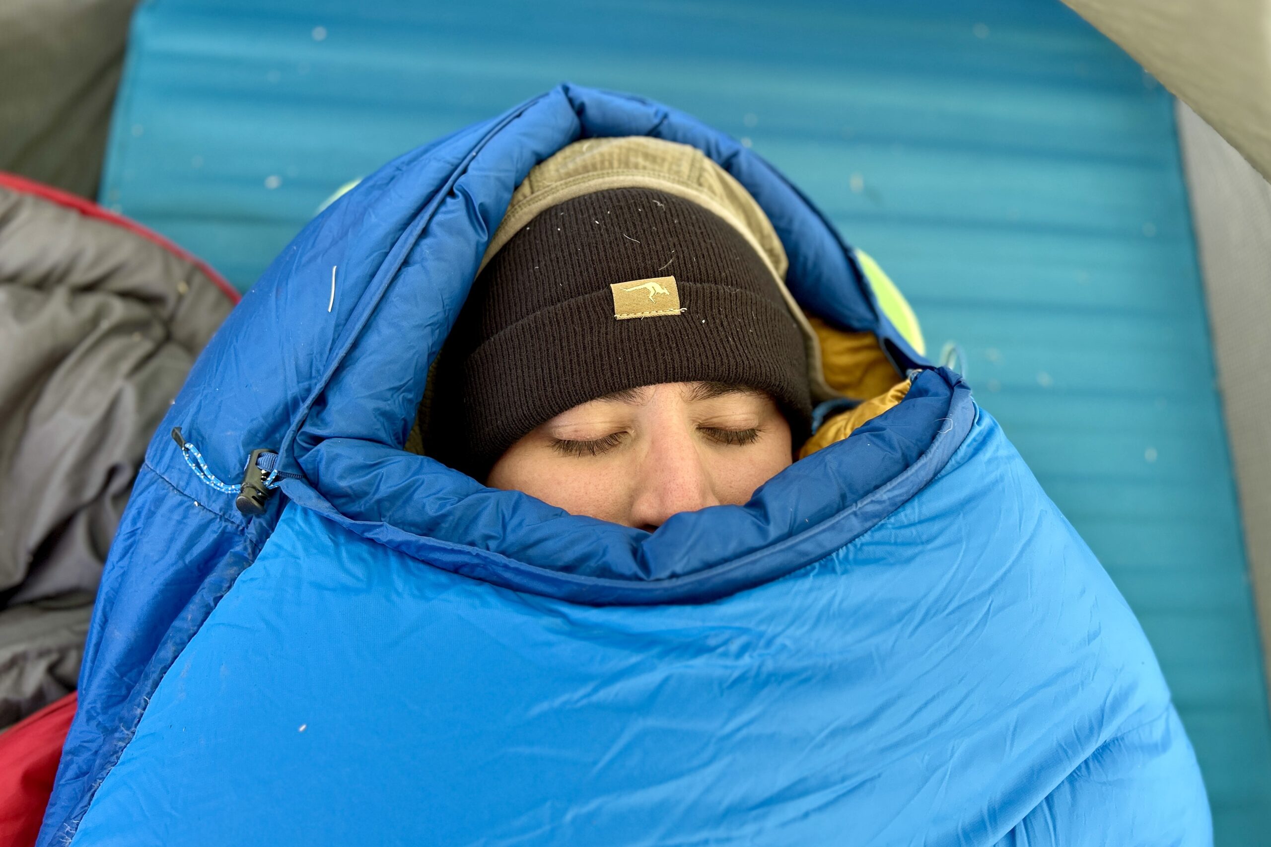 Close-up image of a person's face barely peeking out of a closed mummy bag with the mummy hood up and the bag closed to the top.