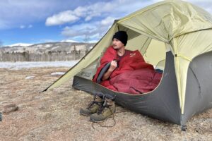A man looks out of a tent into a snowy, mountainous landscape with a sleeping bag wrapped over his shoulders.