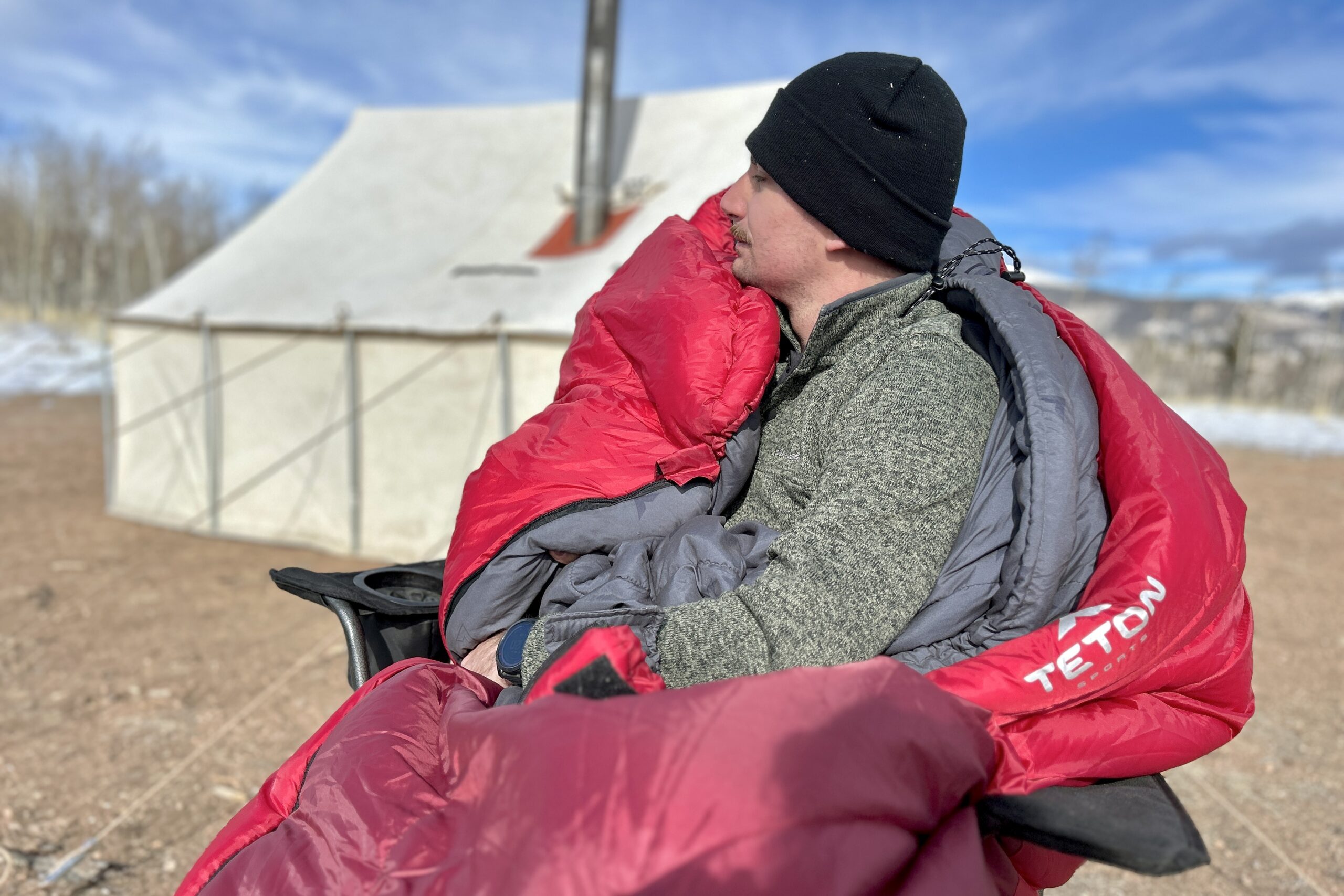 A man sits in a chair in a big, cushy sleeping bag with a canvas tent and snowy mountains in the background.