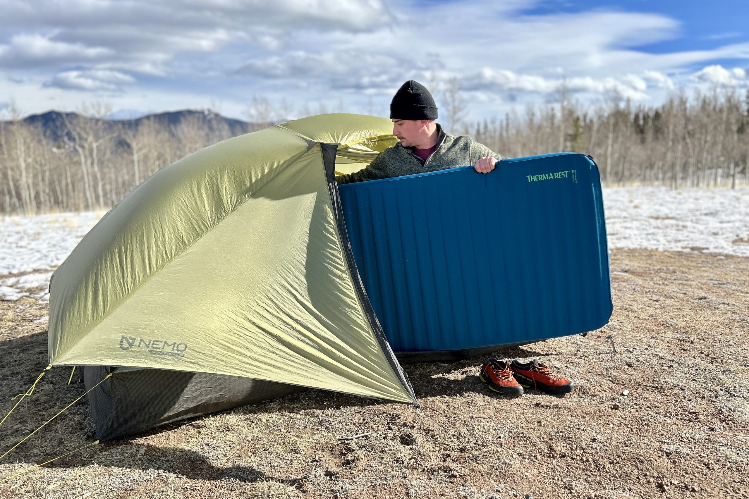 A man puts a wide sleeping mattress into a tent in a snowy landscape.