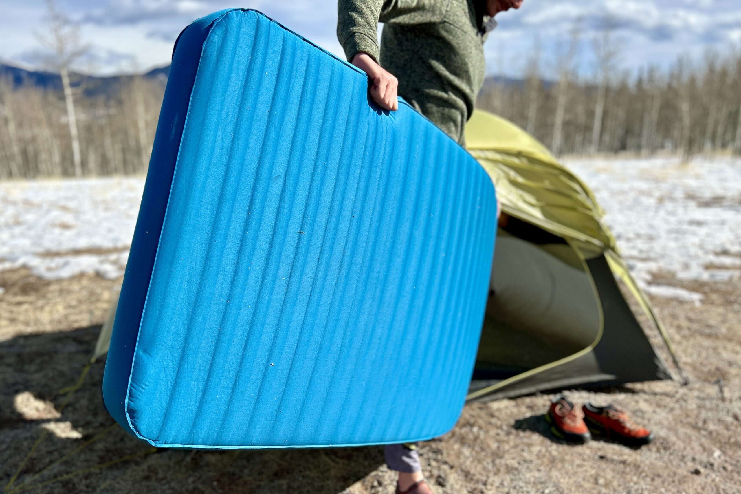 Close-up image of a person loading a wide sleeping mattress into a tent in a snowy landscape.