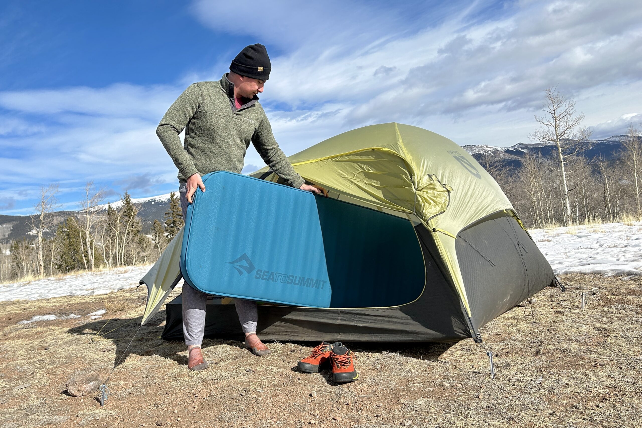 A man loads a camping mattress into a tent in a snowy landscape.