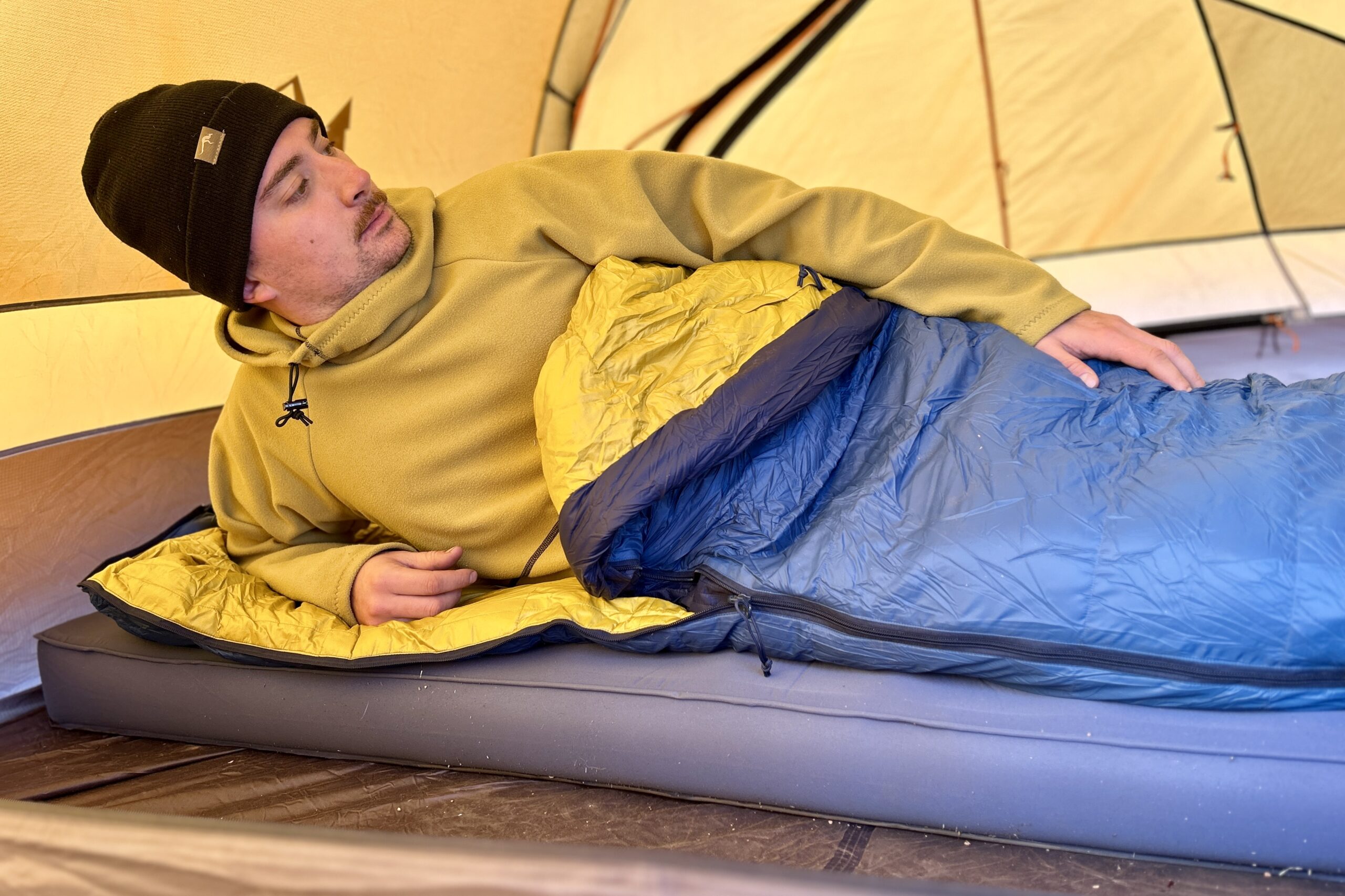 A man sits up on one arm in a a sleeping bag on a camping mattress in a tent.